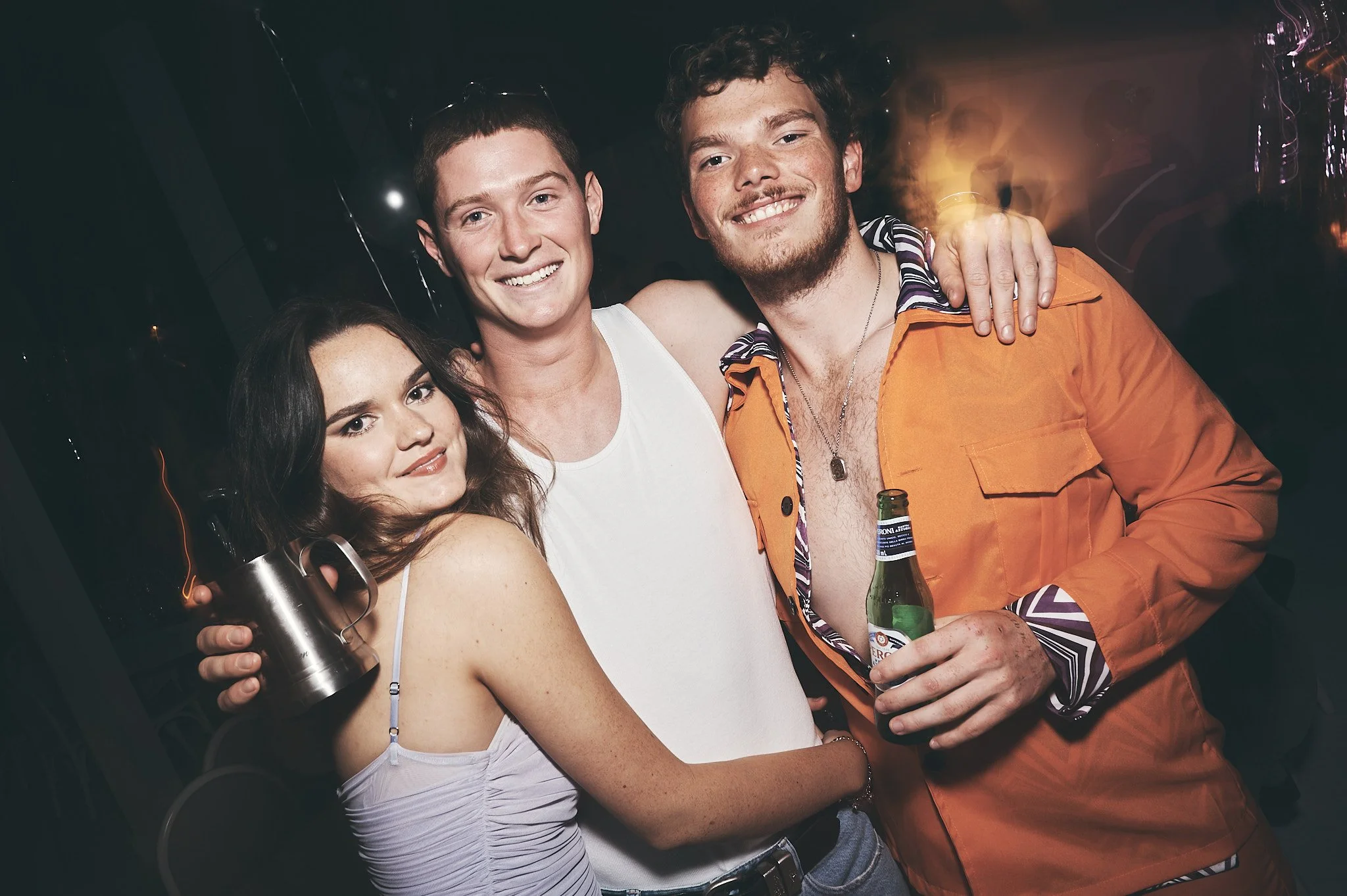 Three young adults posing together at a party or bar, with the woman holding a drink, and one man holding a beer, all smiling at the camera.