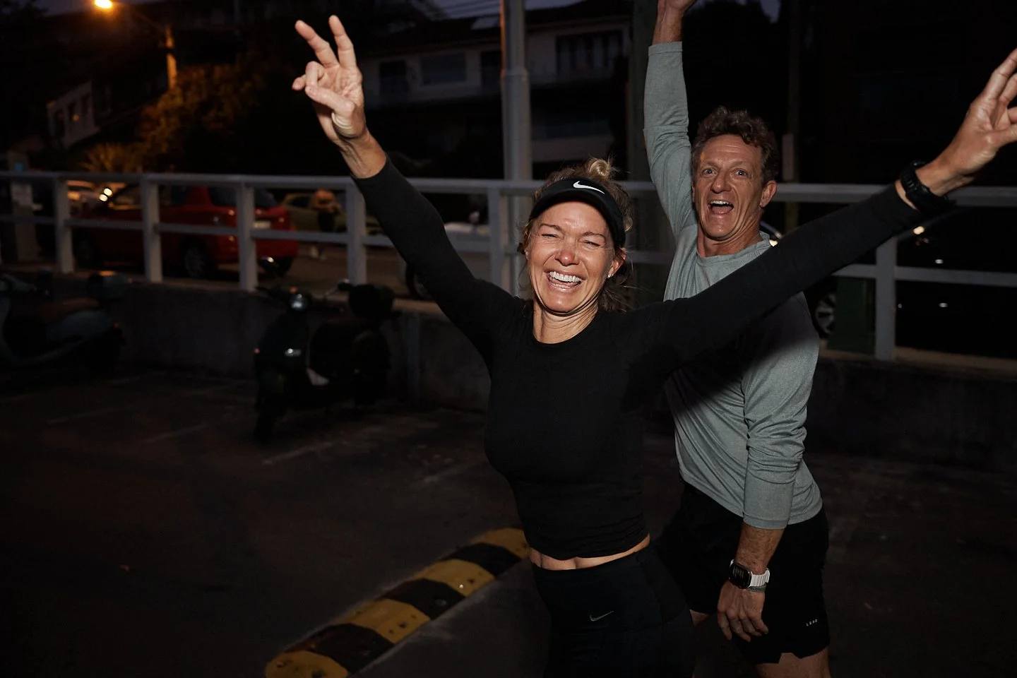 Two people celebrating after running outdoors at dusk, both smiling and raising their arms in triumph, with parking lot and buildings in the background.