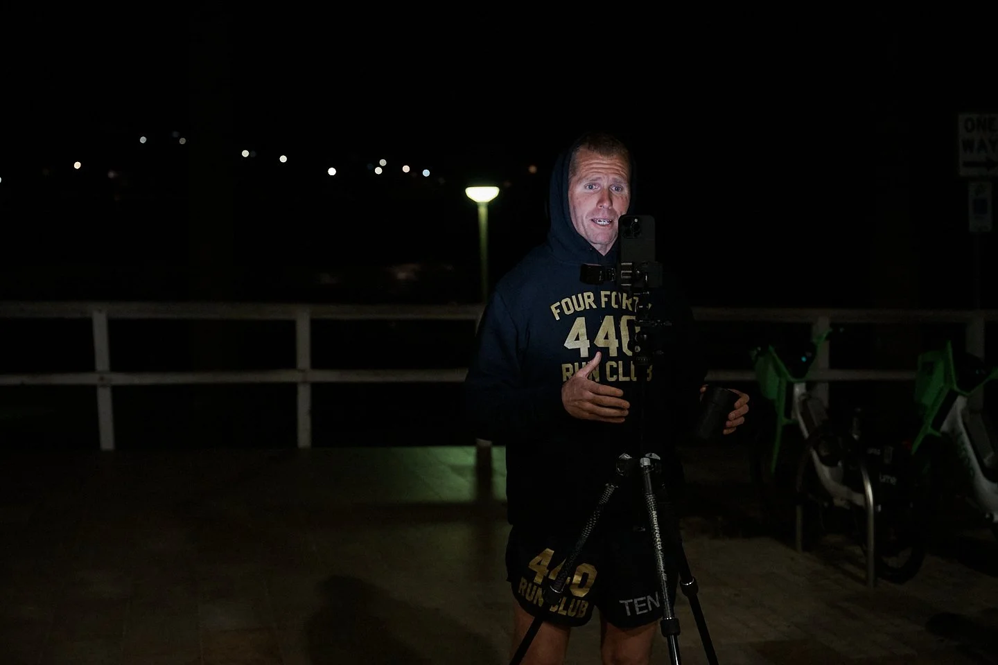 A man in dark athletic wear standing outside at night holding a camera on a tripod, with a lit streetlamp and bicycles in the background.