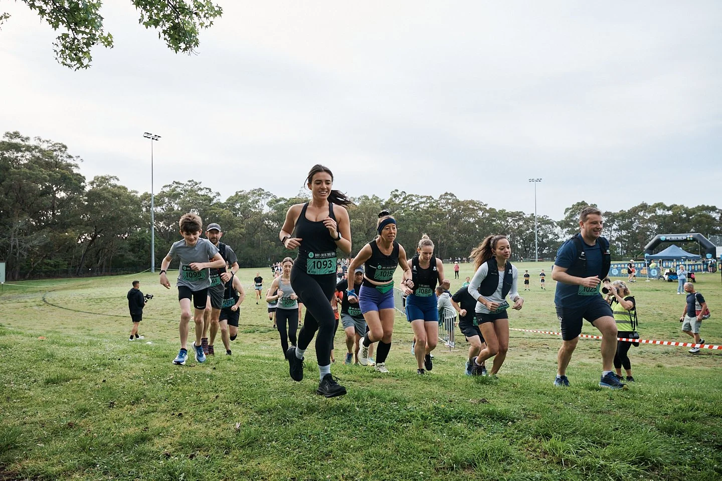 People participating in a race on a grassy field with trees and a sports facility in the background.