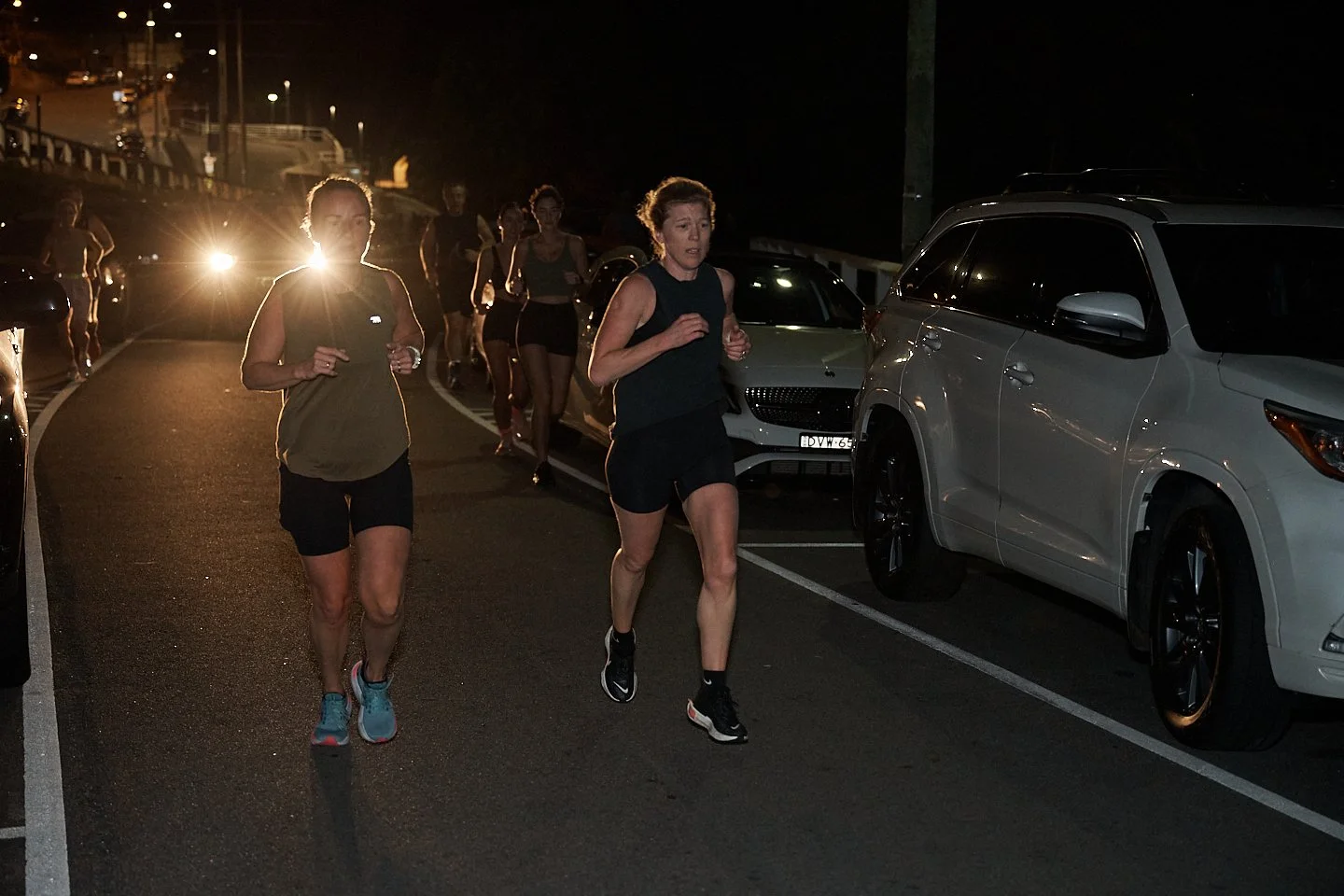 A group of people running on a street at night, with headlights in the background and parked cars on the sides.