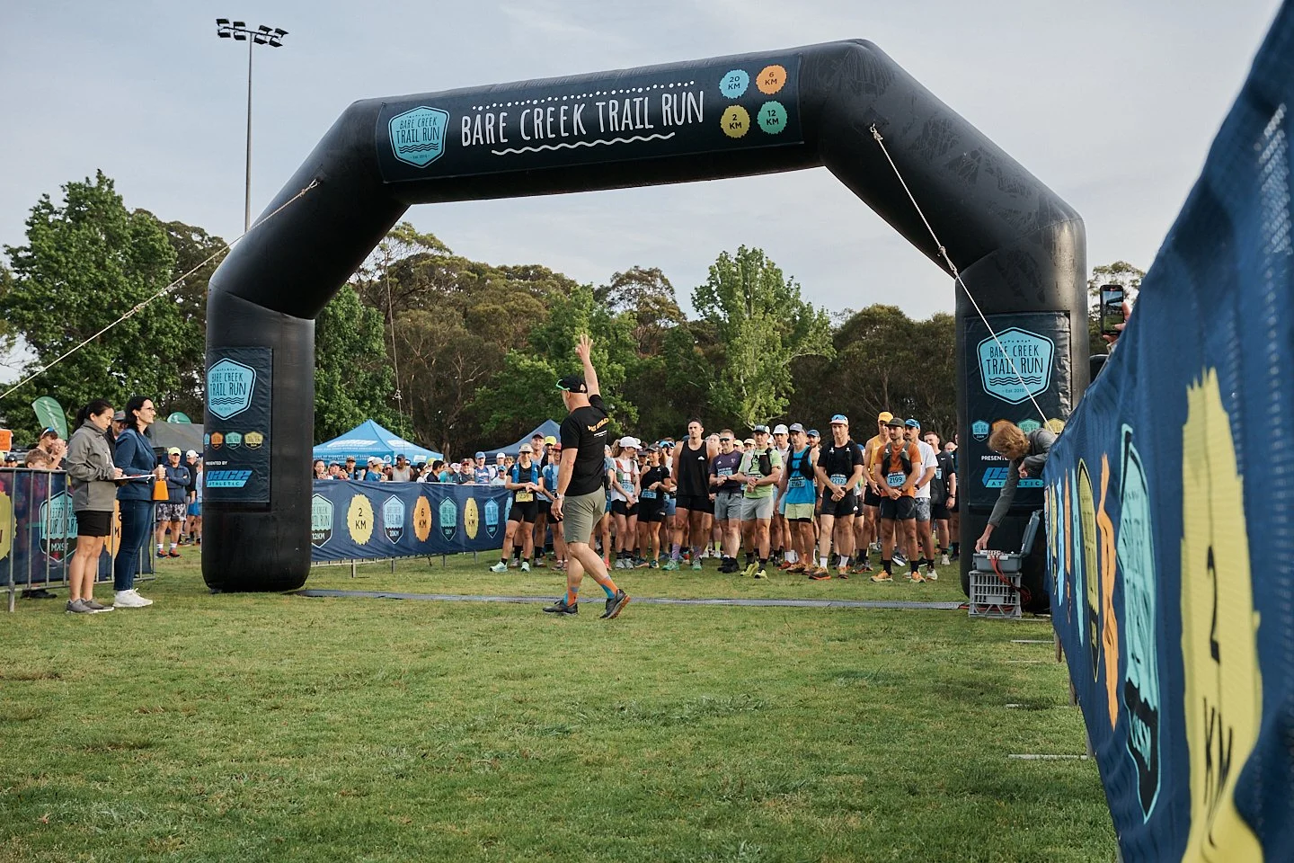 Start line of a trail running race under an inflatable archway that reads 'Bare Creek Trail Run,' with many participants gathered behind the barrier, some wearing running gear and bibs, in a park with trees in the background.