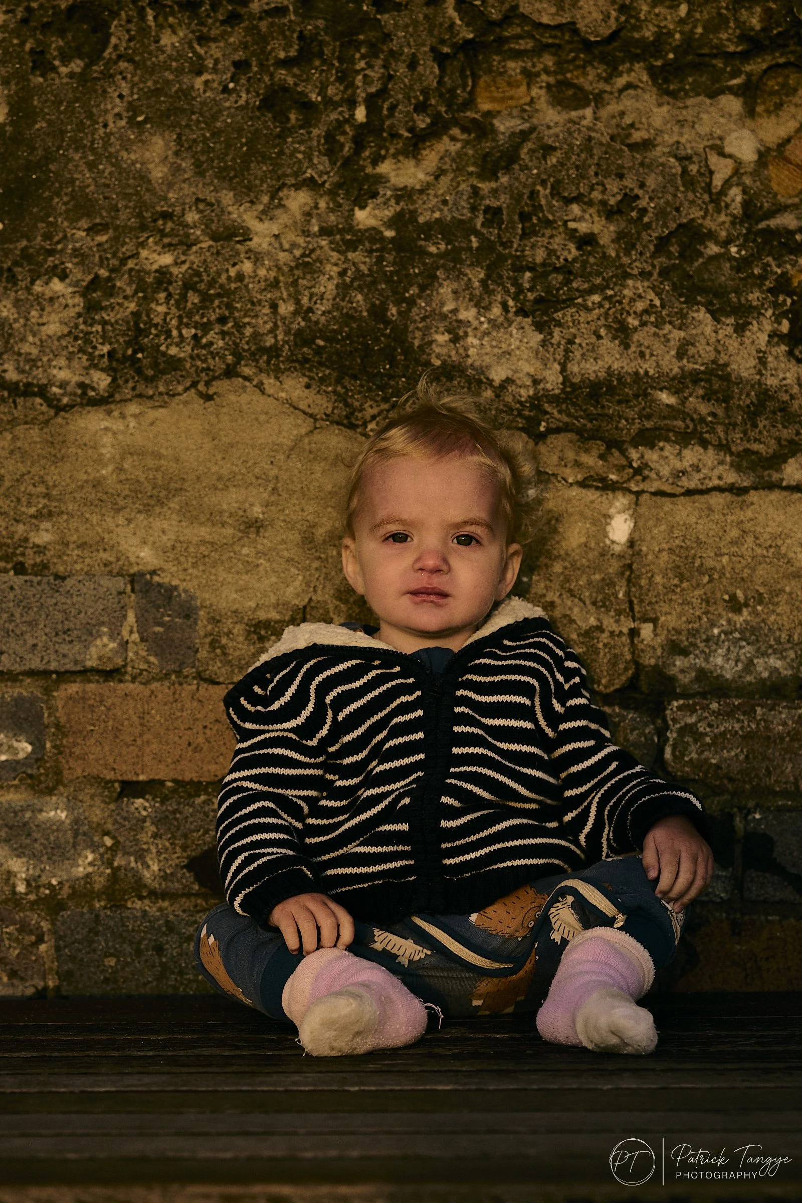 a 2 year old sits leaning against the textured wall at Coogee beach and looks straight at the camera