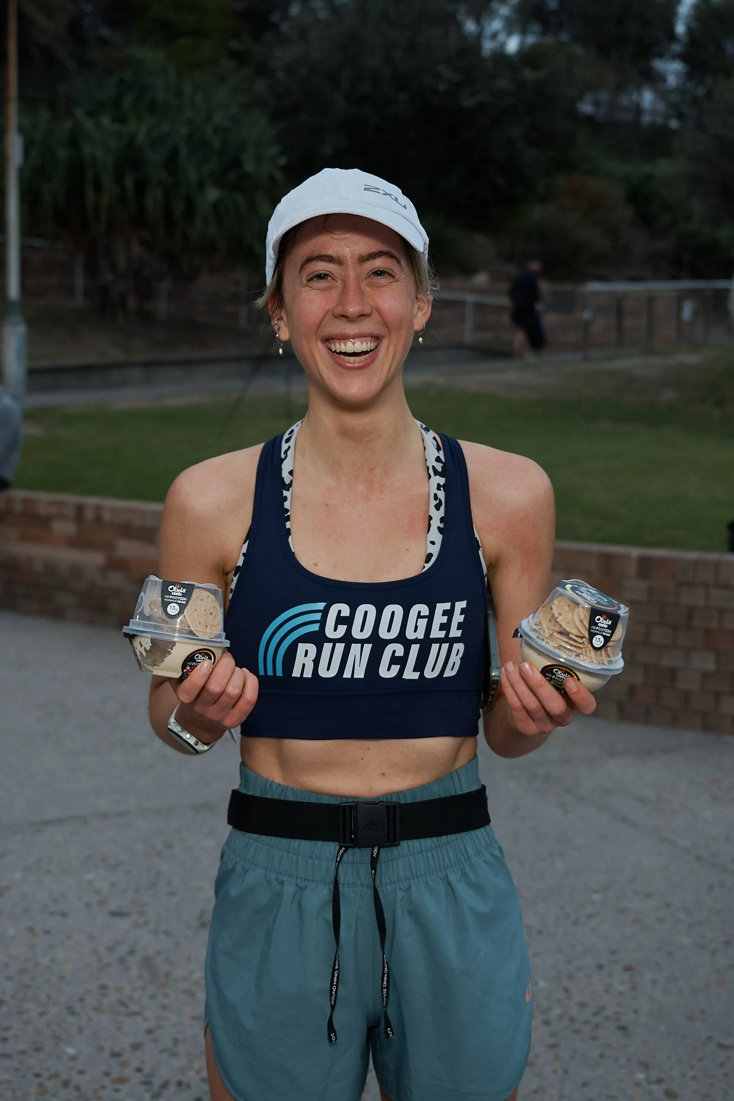 A smiling female runner in athletic gear holding two containers of ice cream during a run or race, with a background of trees and a brick wall.