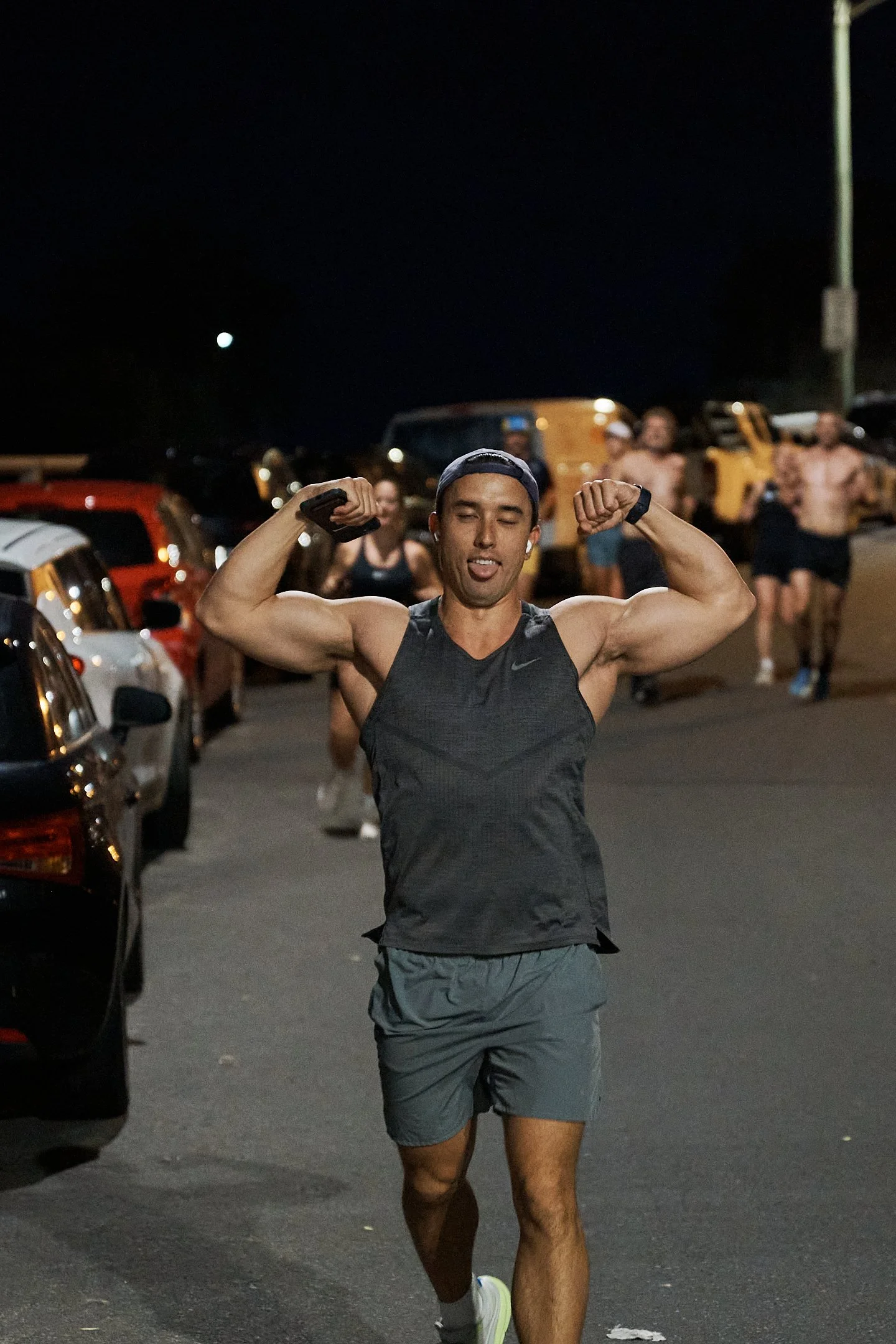 A man running at night, smiling and flexing his biceps, with other runners in the background on a parking lot street.