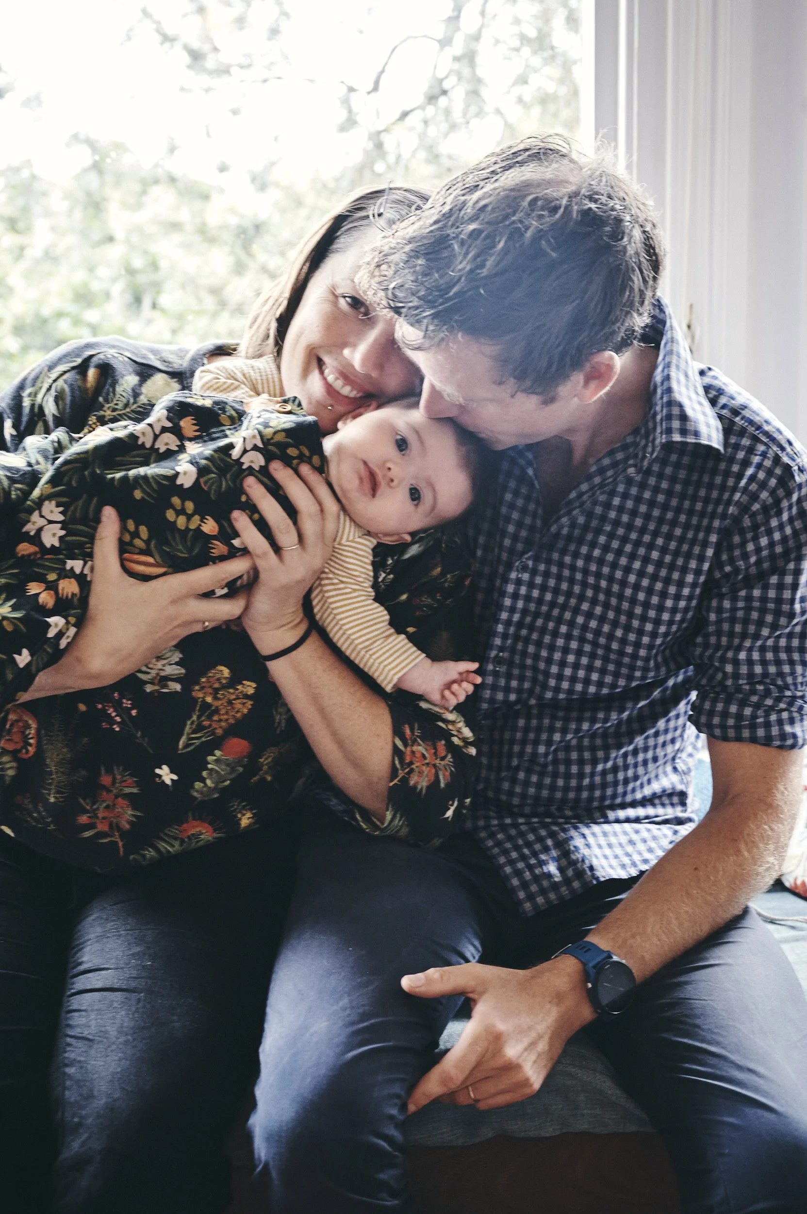 A family of three sitting near a window with natural light. The woman is holding a baby, and the man is leaning in close, both smiling and showing affection to the baby. The scene is warm and intimate.