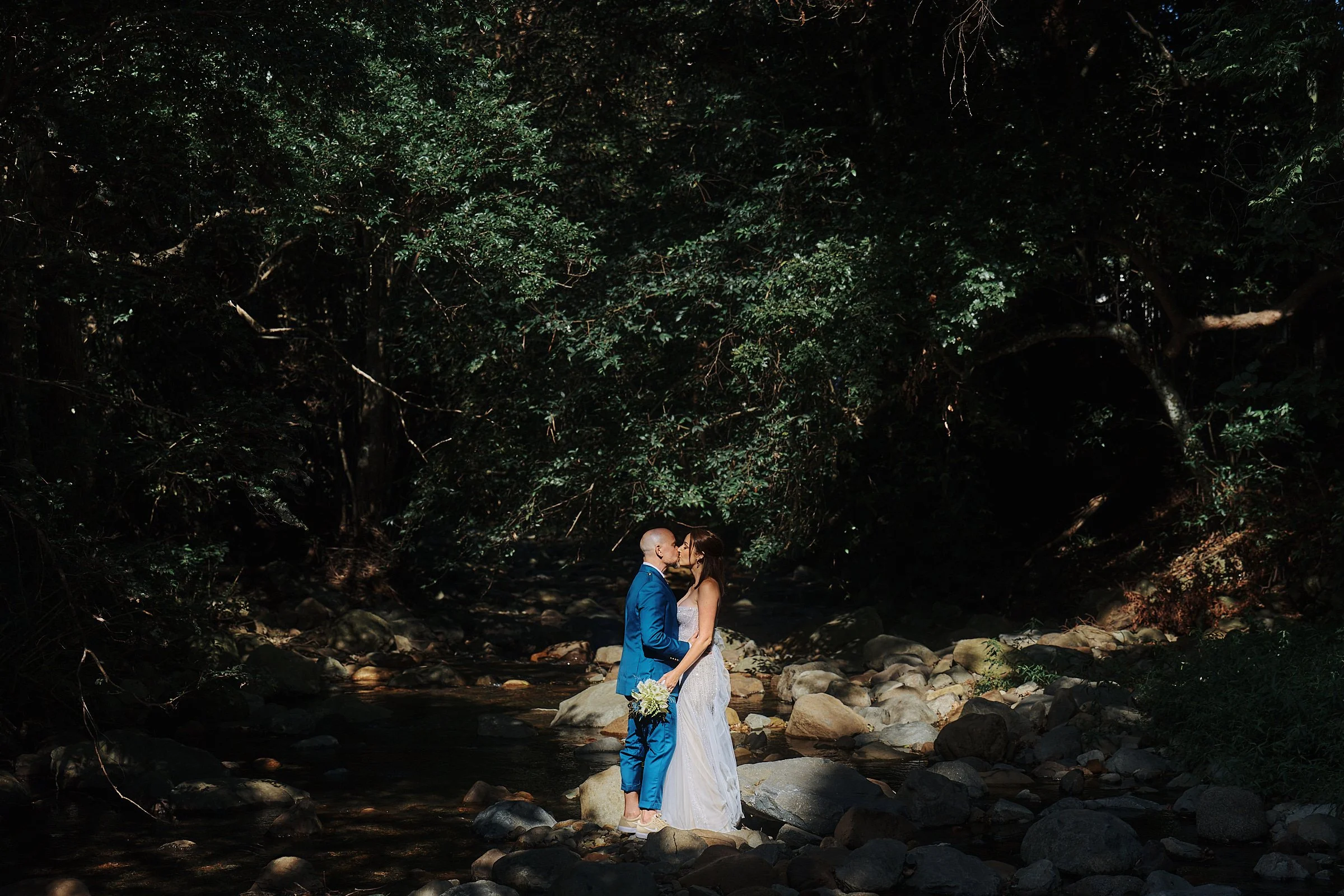 a newly married couple stand in a creek, lit by dappled sunlight, and share a kiss
