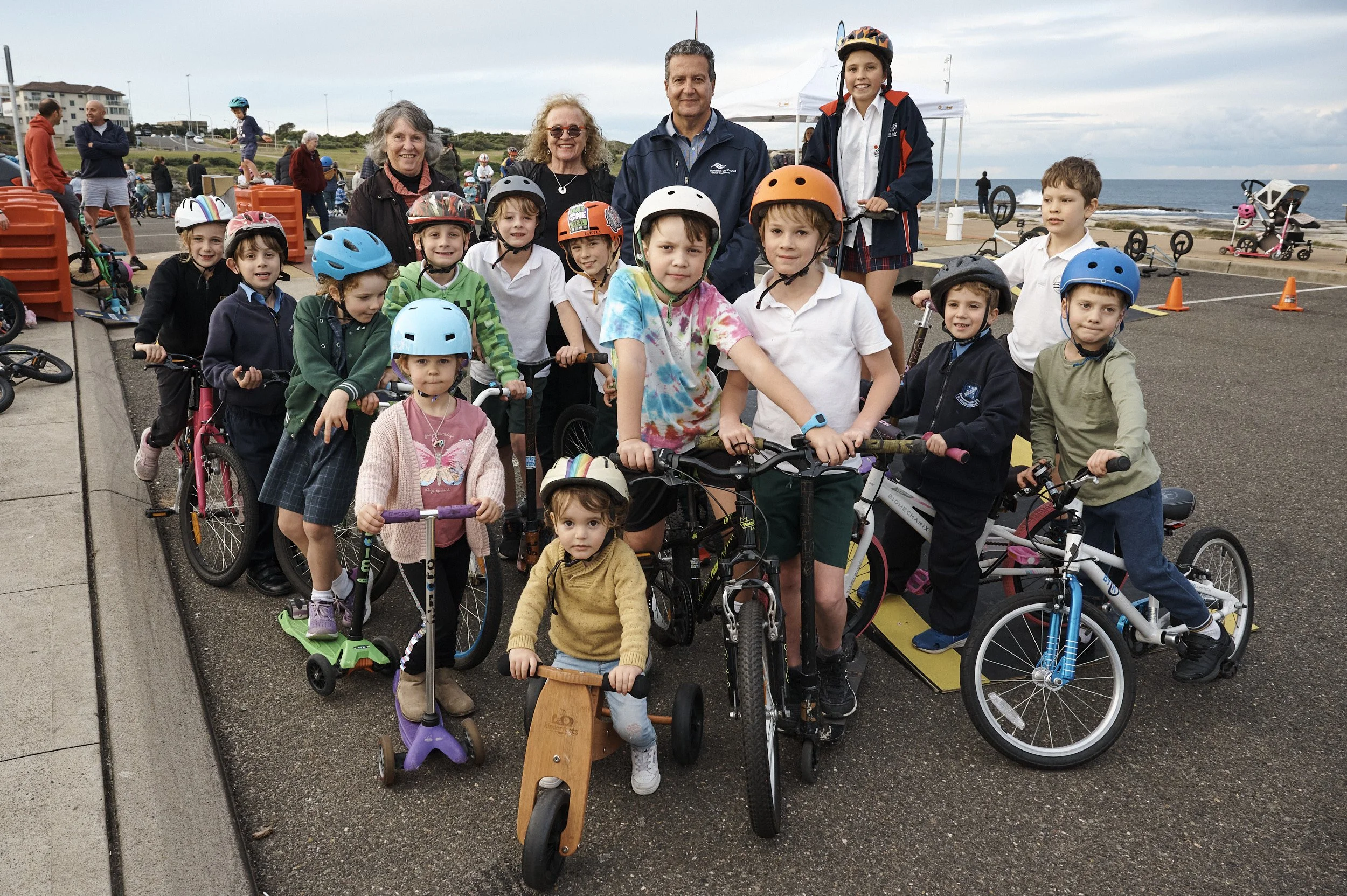 Group of children with helmets and scooters, bikes, and skateboards, along with a few adults, gathered outdoors near a beach with a cloudy sky.