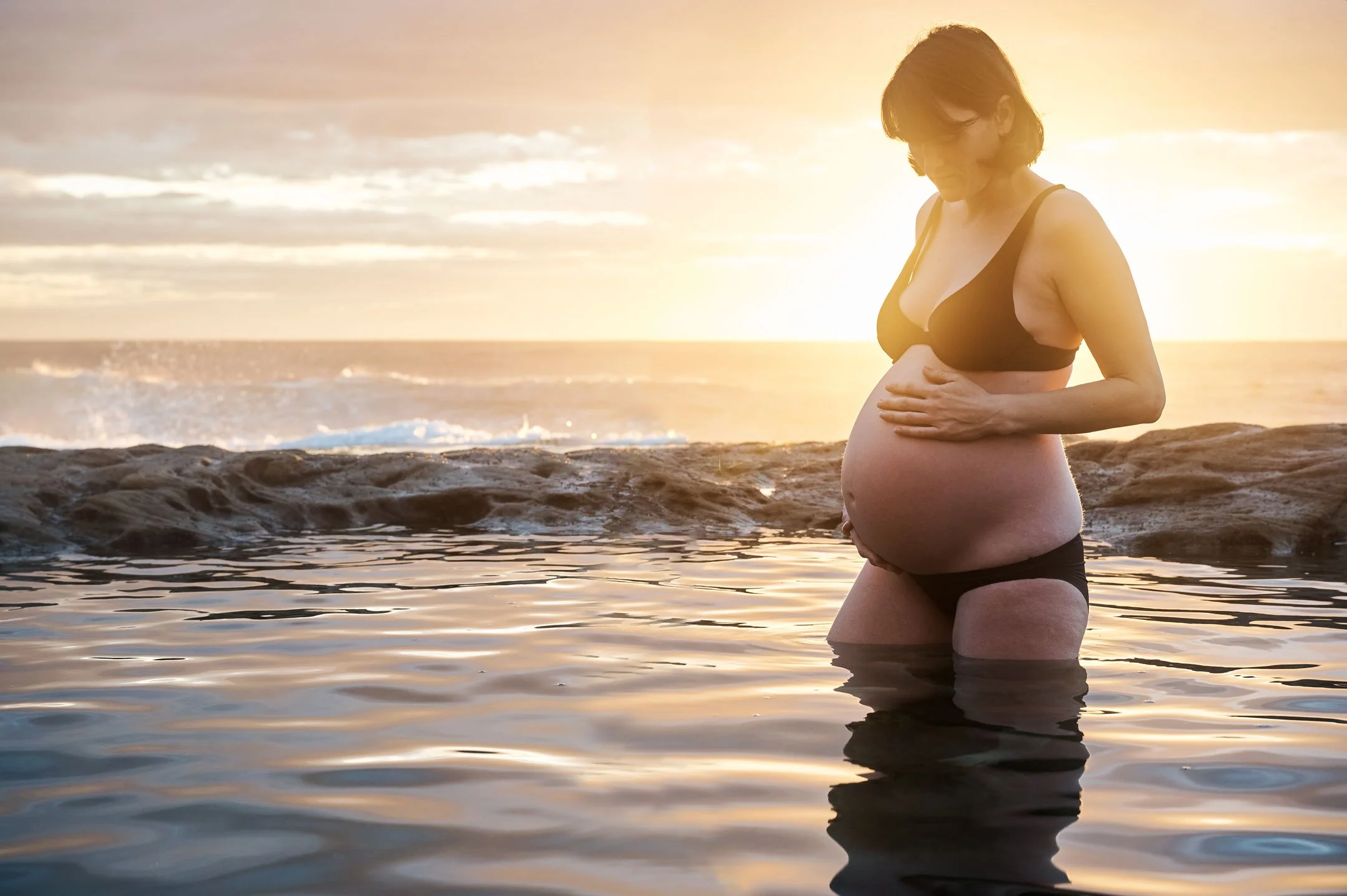 a pregnant woman stands in the water during sunrise at Ivor Rowe Rock Pool in Coogee