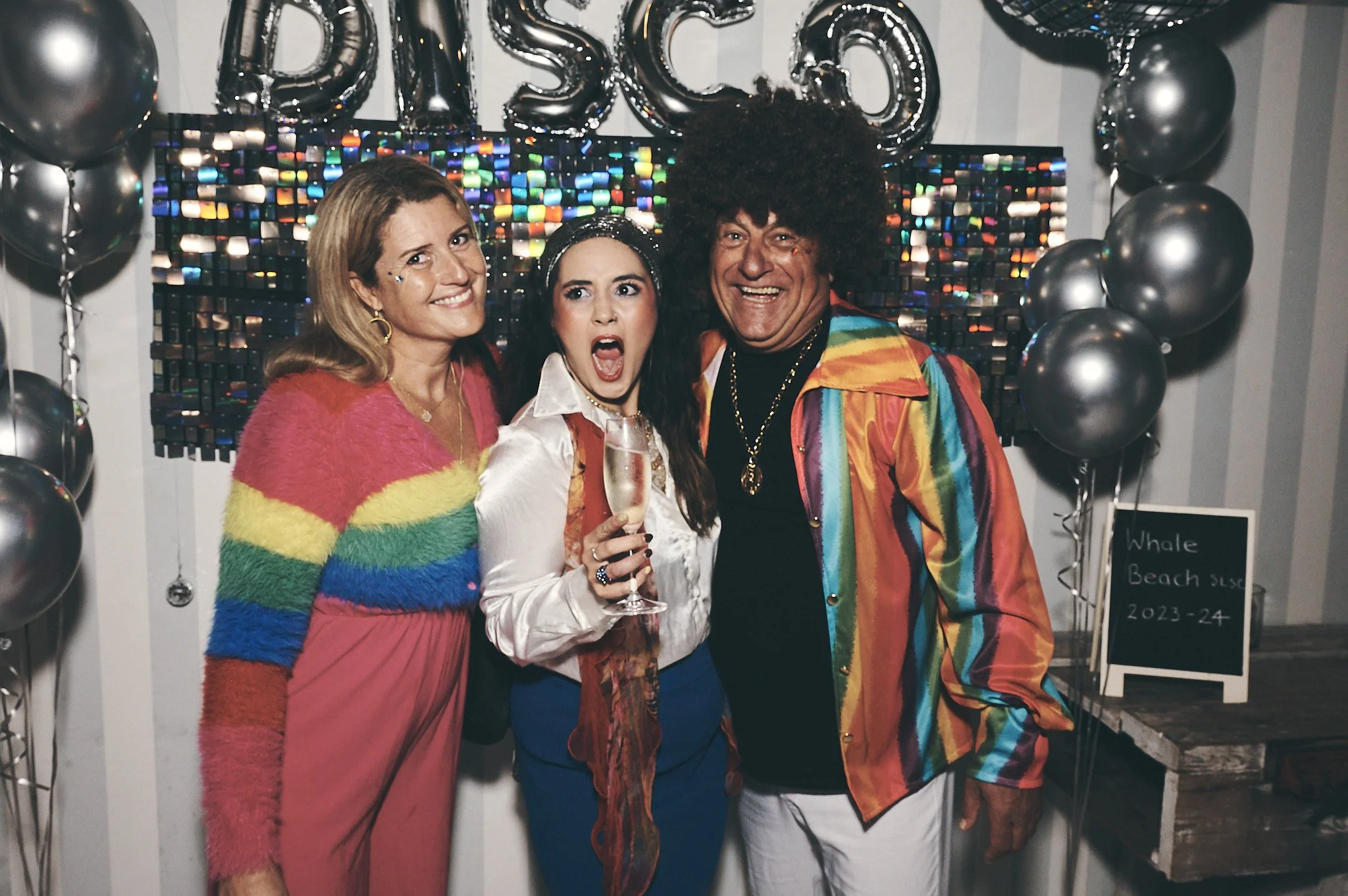 Three people celebrating at a party; they are wearing colorful, retro-style clothing and are standing in front of a decorated wall with balloons, a silver 'DISCO' balloon, and a sign that reads 'Whole Beach 2023-24'.