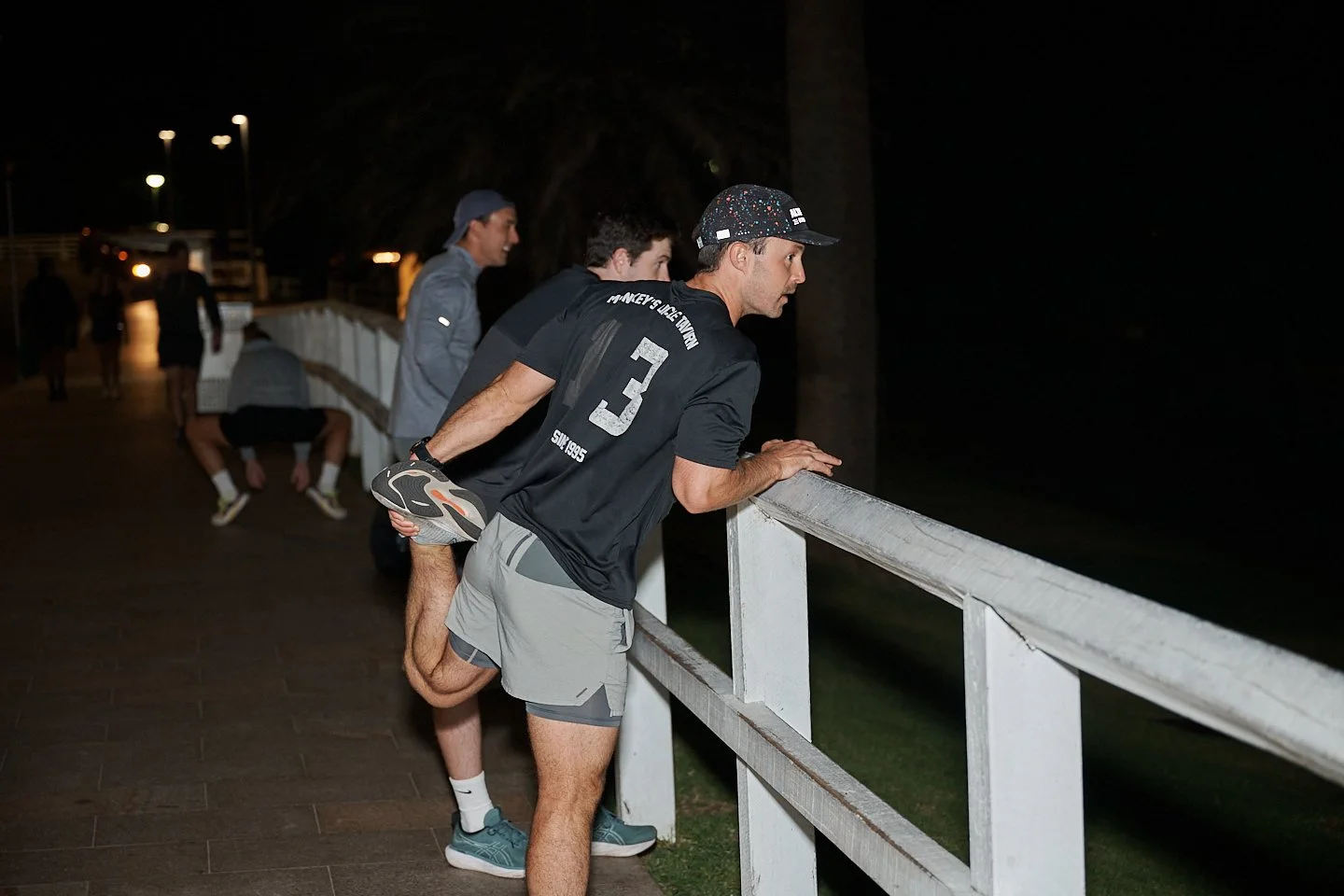 A man stretches his leg behind him while leaning on a white fence at night, watching something in the darkness. Other people in athletic gear sit or stand along the fence nearby.