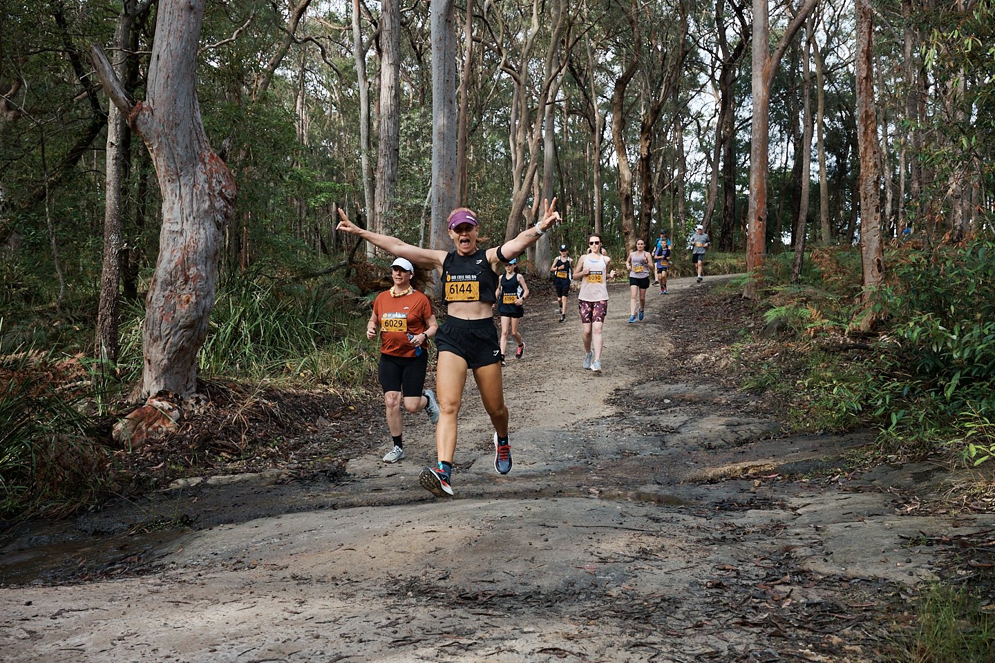 A group of runners participating in a race on a wooded trail, with one woman in the front making peace signs and smiling, wearing a black top, shorts, and a purple visor.
