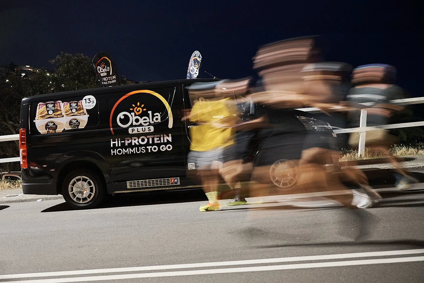 Nighttime street scene with a black van advertizing Obela high-protein hummus, featuring a logo and product images, while a group of people in motion blur walk past the van.