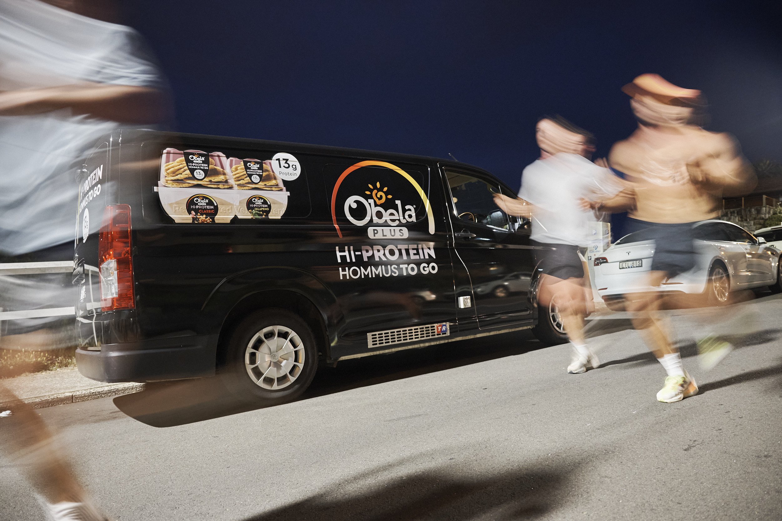 People running past a black Obela Plus delivery van advertising high-protein hummus to go, with a night sky in the background.