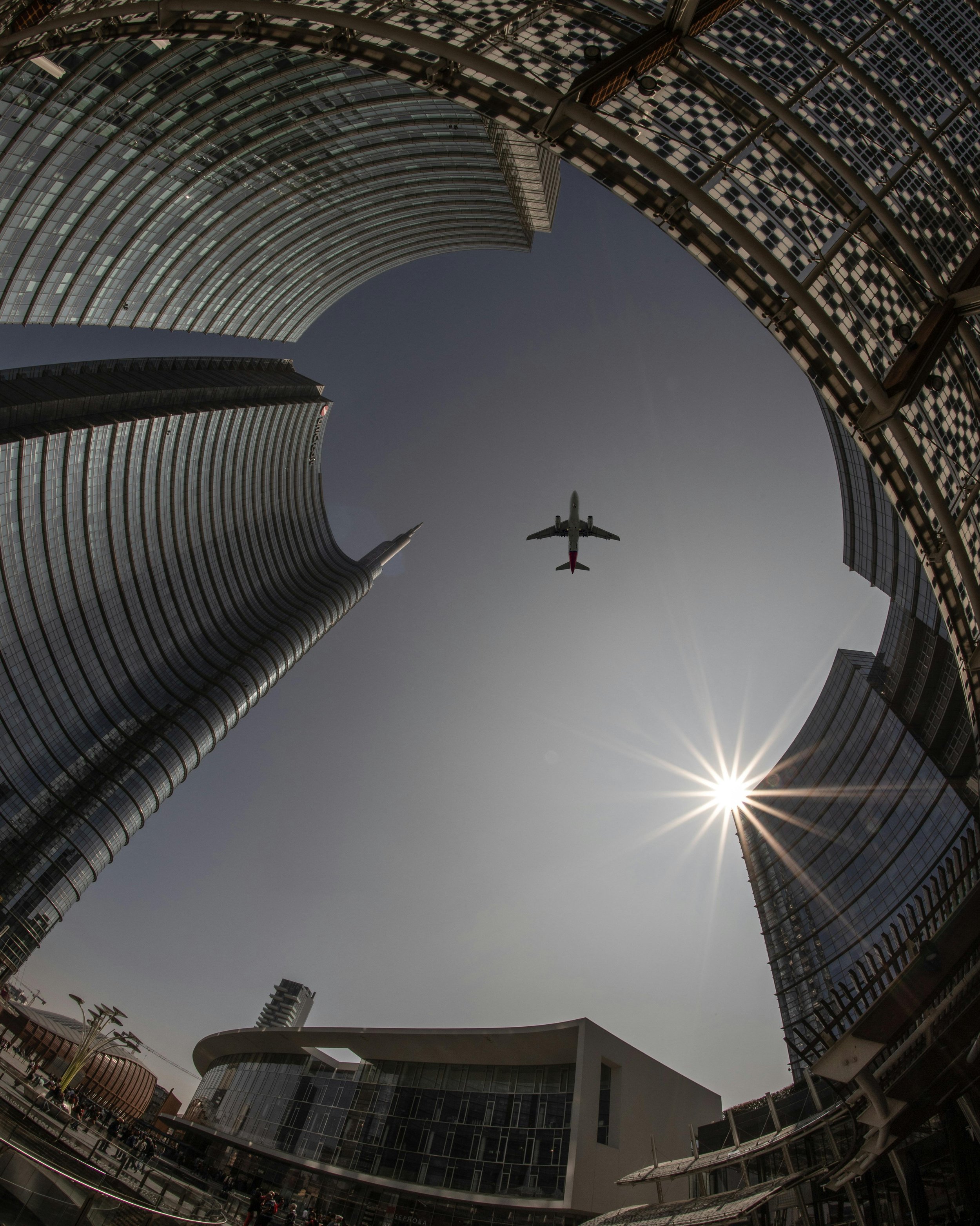 A low-angle view of skyscrapers in an urban area with an airplane flying overhead against a clear sky and the sun shining brightly.