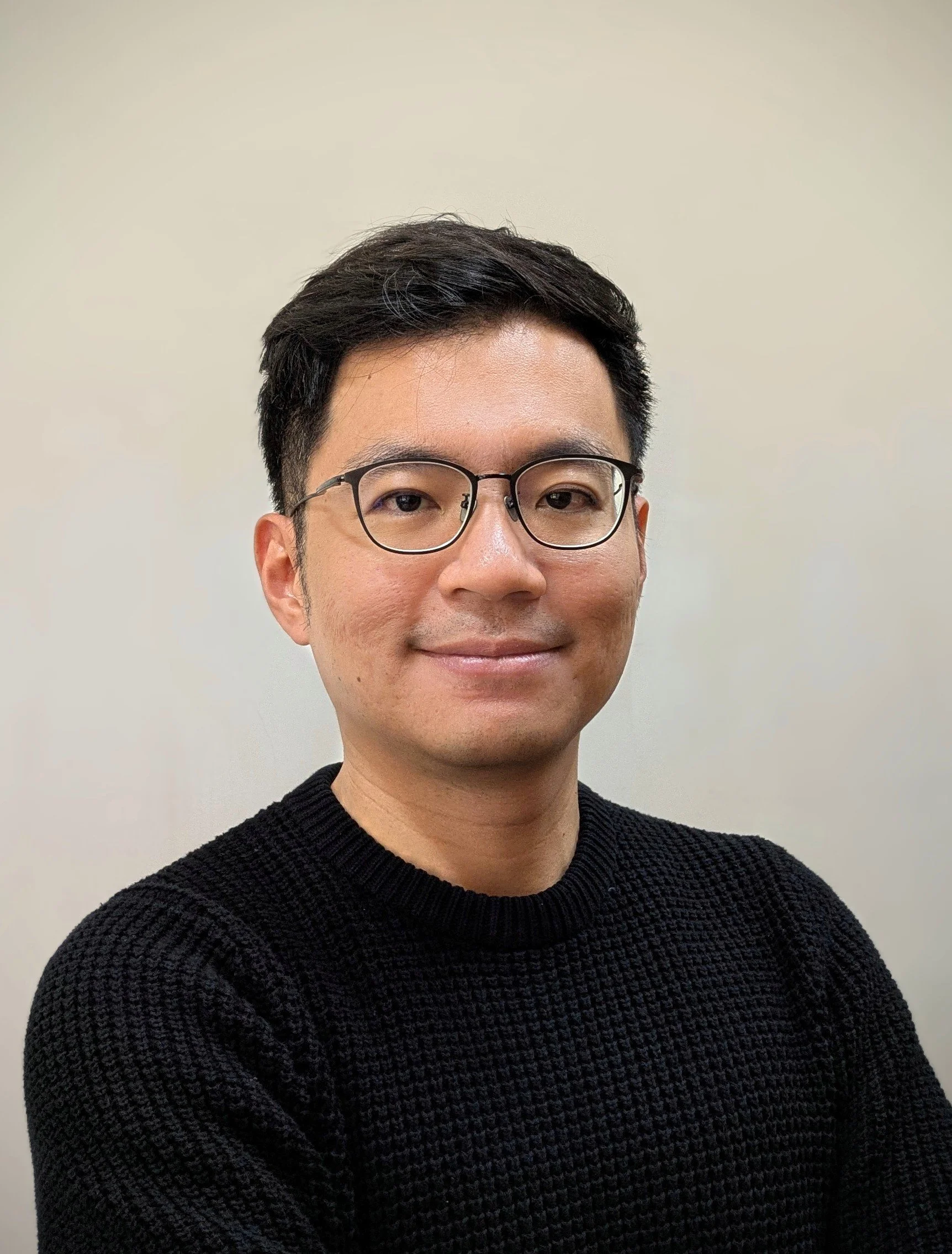 Portrait of a man with short black hair, glasses, wearing a black sweater, smiling, against a plain light-colored background.