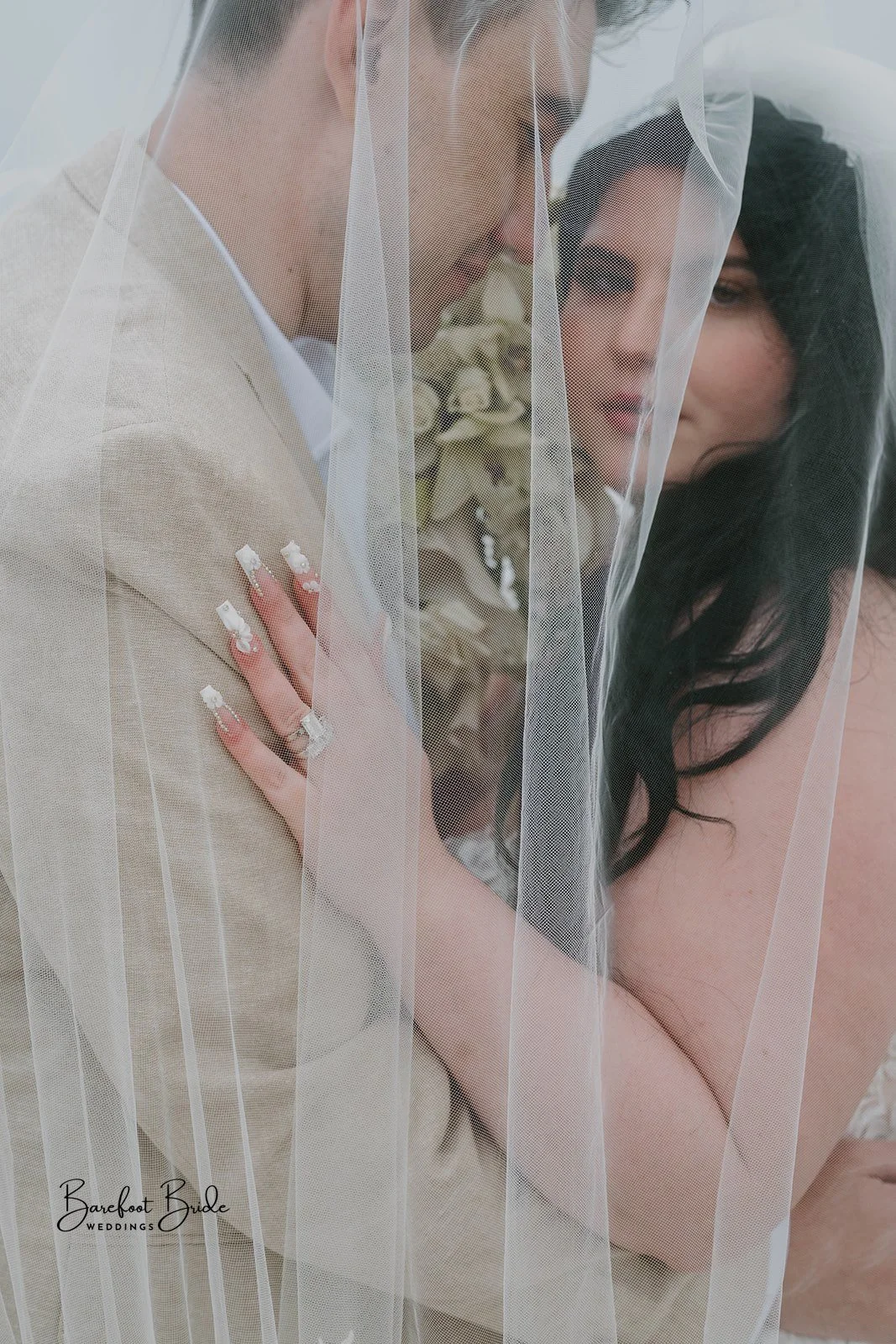 A bride and groom cover with a sheer veil, with the bride's hand resting on the groom's shoulder, showing rings and decorated nails, close-up shot.