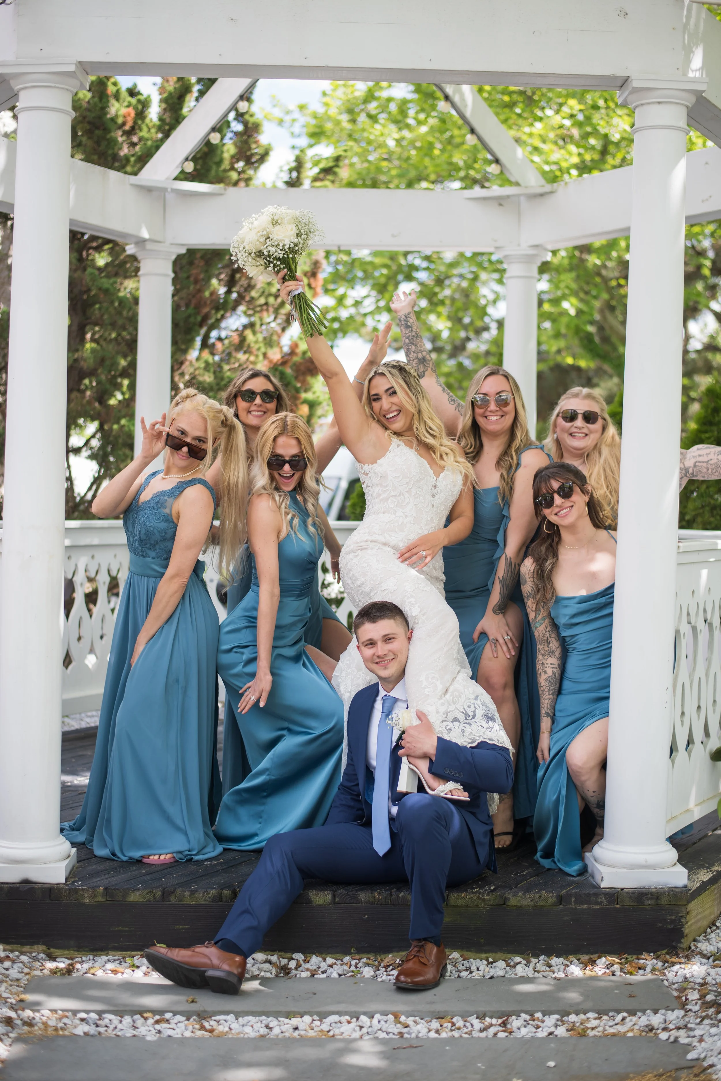 A group of women in blue dresses and a man in a suit gathered around a bride in a white wedding dress, at a wedding celebration, outdoors under a white pavilion with trees in the background. The women and man are smiling, with some wearing sunglasses