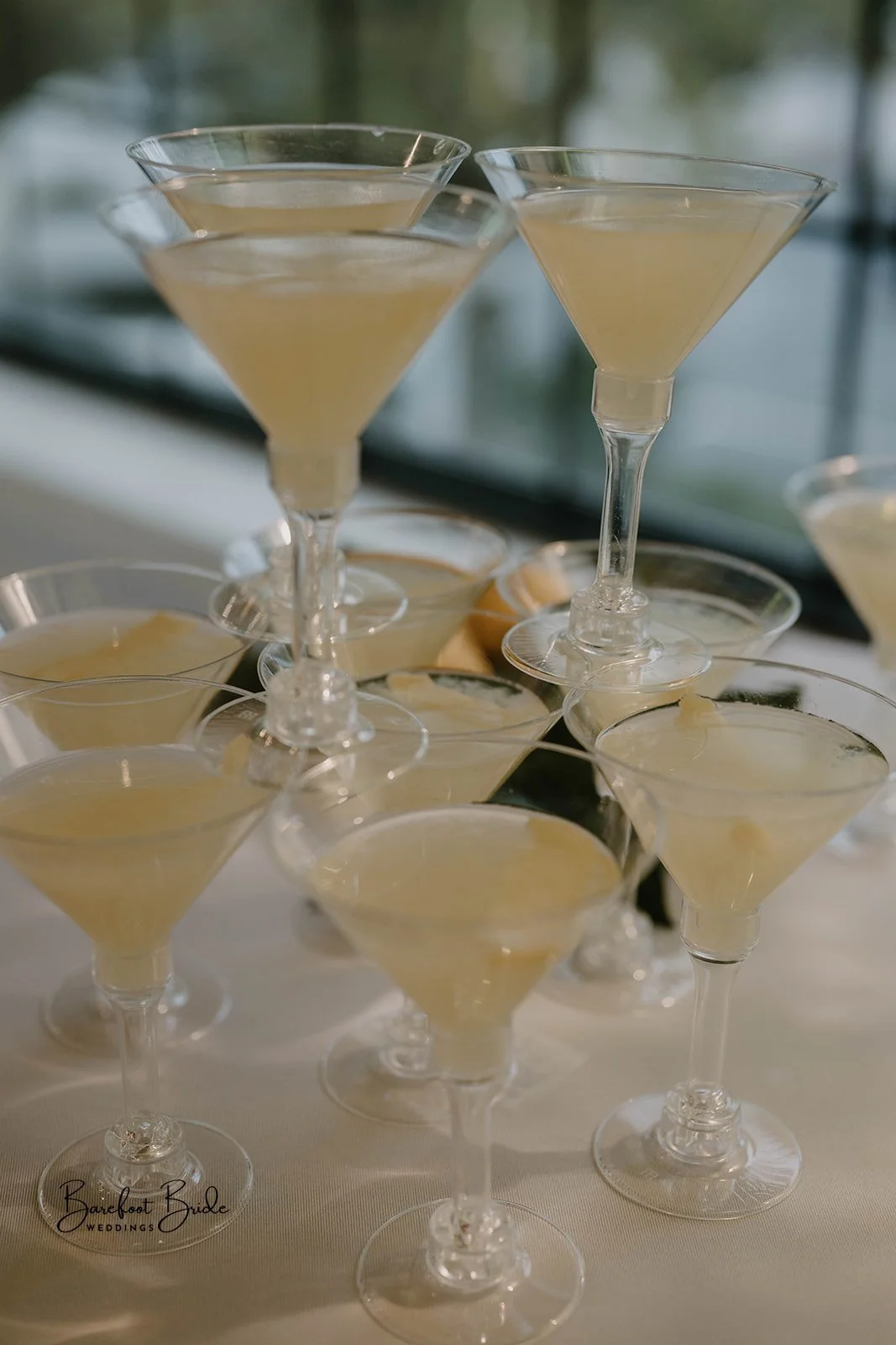 Multiple cocktails in martini glasses stacked on a table, some garnished with lemon slices, with a blurred background of windows.
