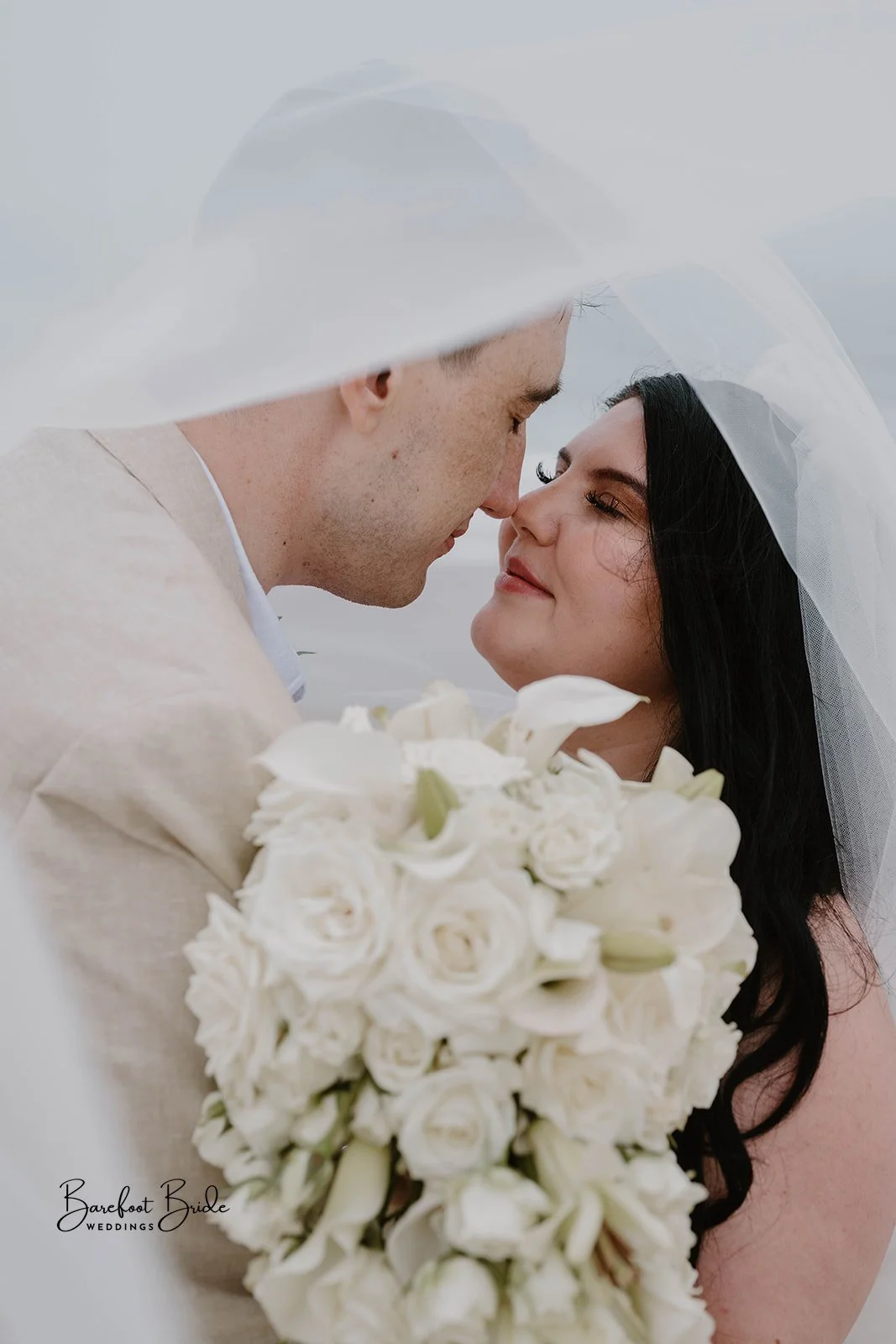 A bride and groom sharing a close moment, with their foreheads touching and eyes closed, holding a large bouquet of white flowers, during their wedding.