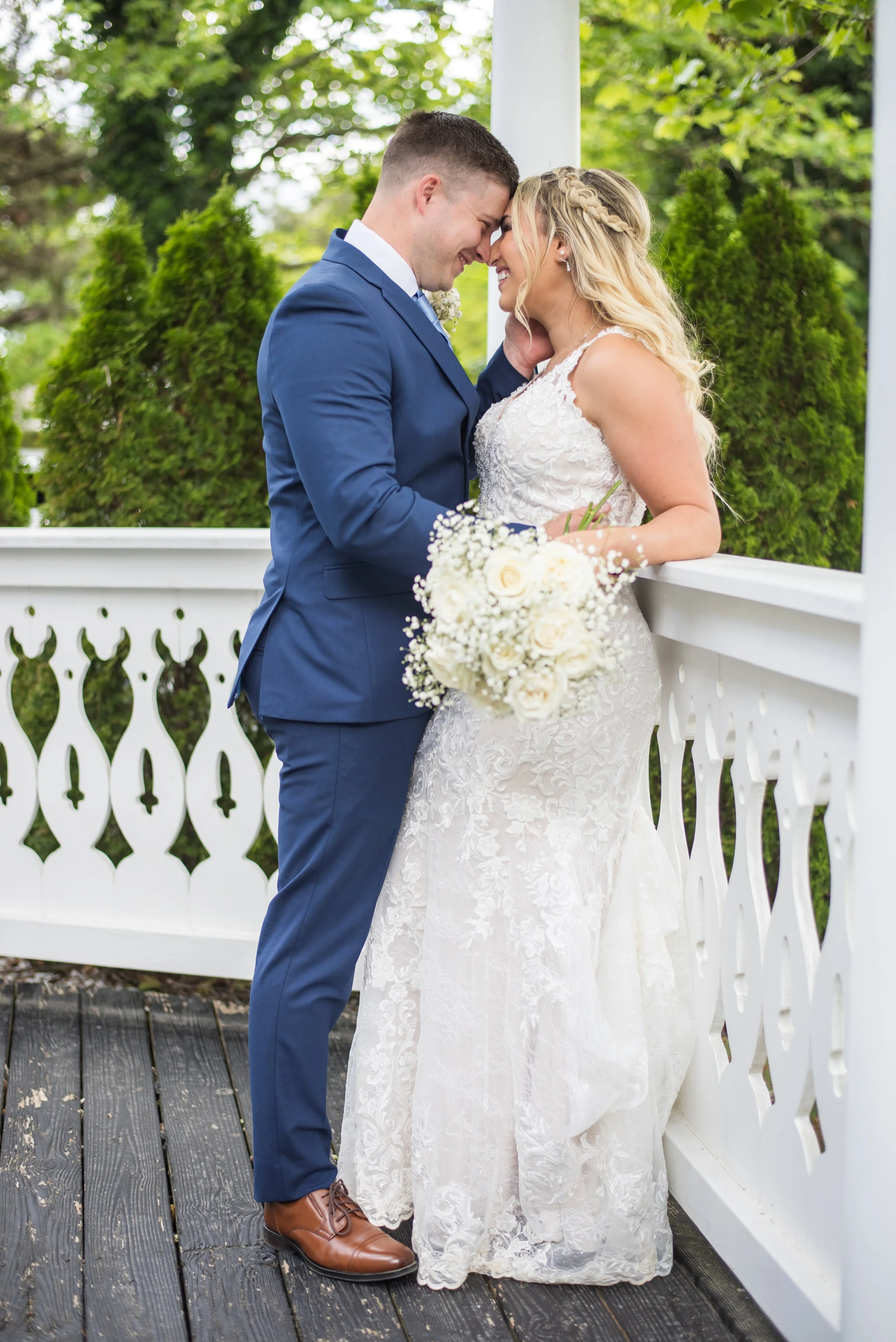 A newlywed couple on a wooden bridge, close together with foreheads touching, smiling, outdoors with green foliage in the background. The groom is wearing a blue suit and brown shoes, holding a bouquet, and the bride is in a white lace wedding dress 