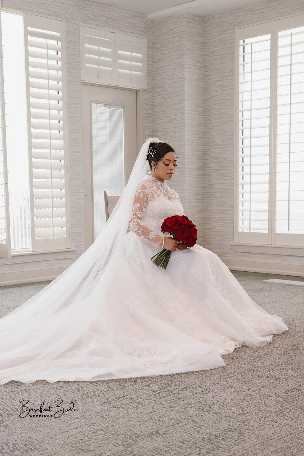 A bride in a white wedding dress with lace sleeves, sitting on a chair in a room with white shutters and light-colored walls, holding a bouquet of red roses, with a long veil flowing on the floor.
