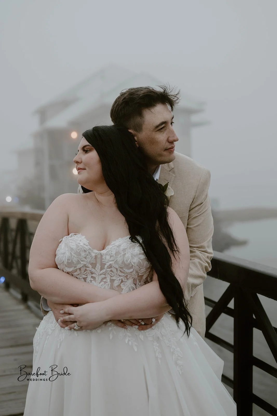 Bride and groom embracing on a foggy dock with a house and ocean in the background.