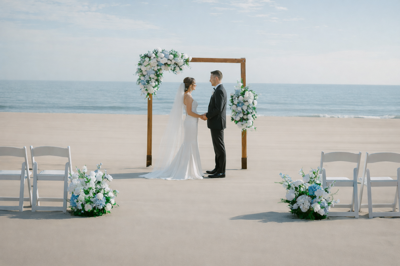 A bride and groom holding hands and facing each other on a beach wedding setup, with an ocean backdrop, decorated with floral arrangements and wooden arch.