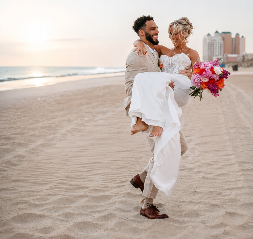 A man in a beige suit carrying a woman in a wedding dress and holding a bouquet of flowers on the beach at sunset.