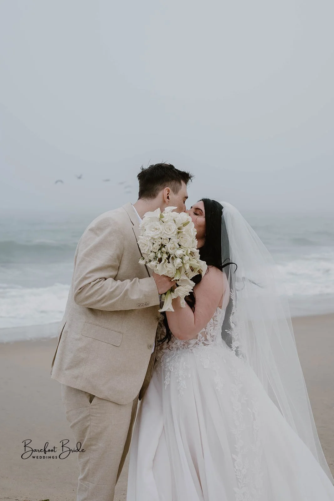 A couple in wedding attire sharing a kiss on the beach, with the groom holding a large bouquet of white roses, and the bride in a lace wedding gown and veil, with the ocean and sky in the background.