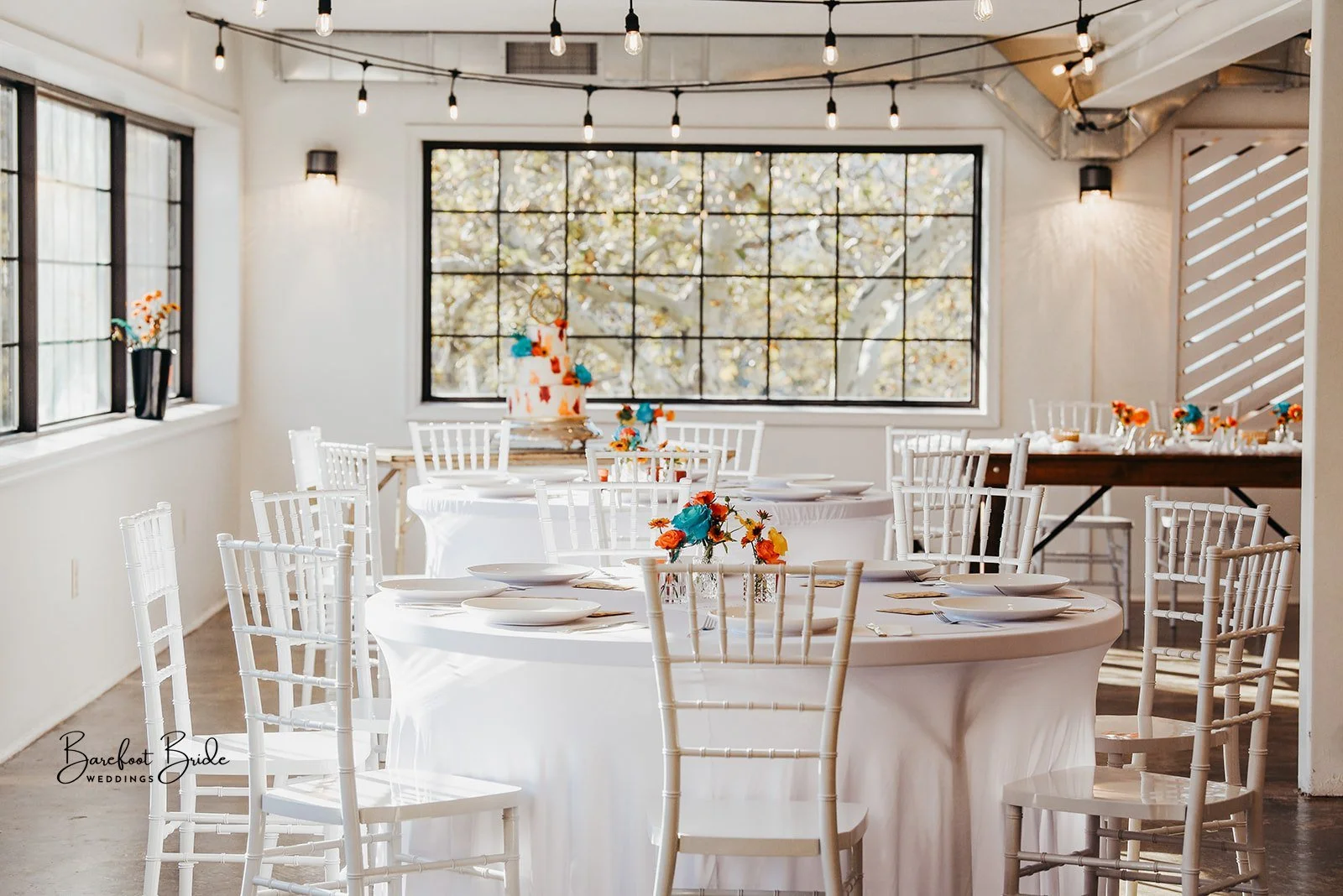 Decorated wedding reception space with round tables draped in white cloths, white chairs, colorful floral centerpieces, a tiered cake with flowers, and large windows letting in natural light.