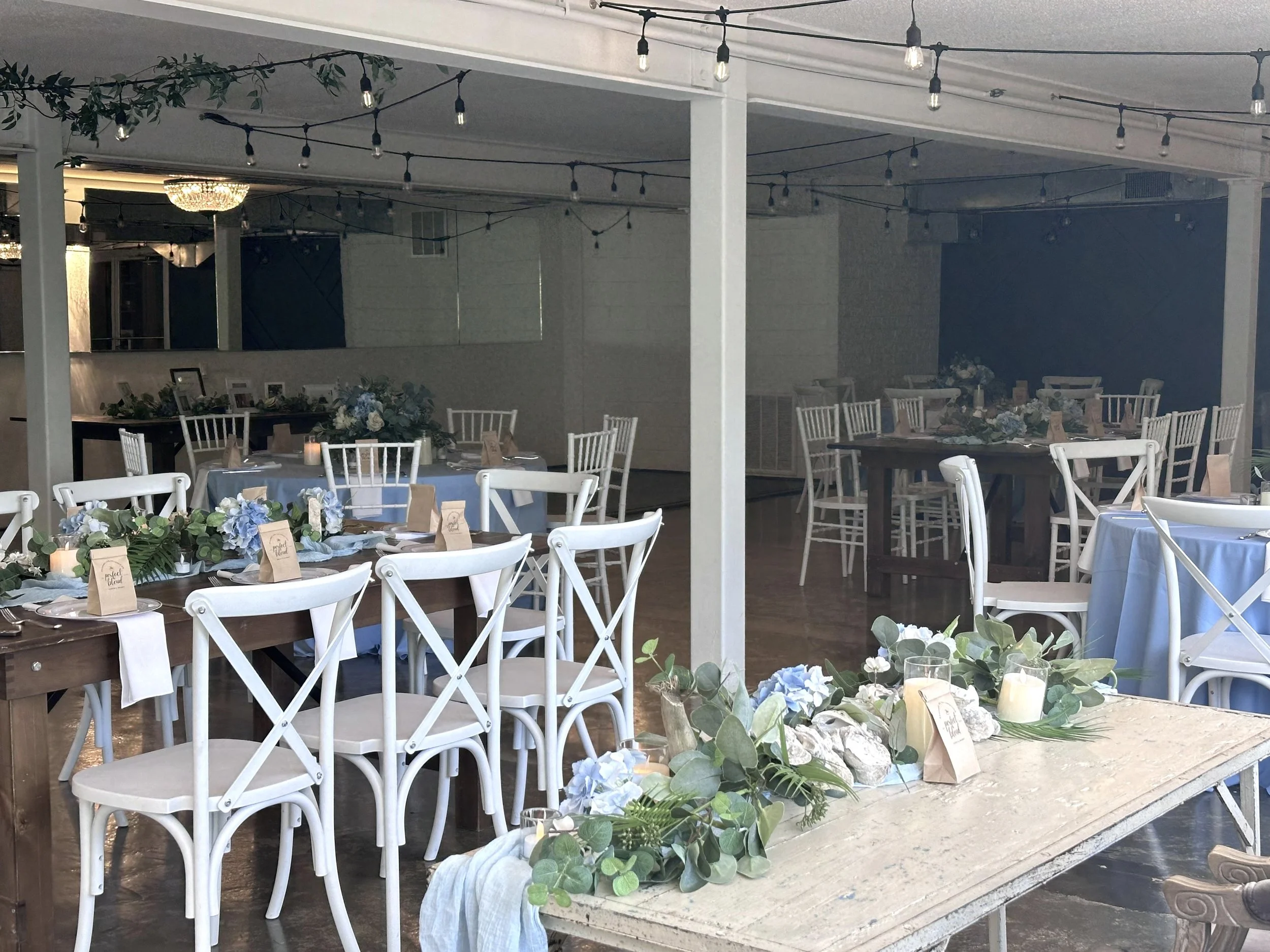 Elegantly decorated indoor event space with round and rectangular tables covered with light blue tablecloths, white chairs, floral centerpieces in soft pastel and greenery, candles, and place cards, set up for a wedding reception or celebration.