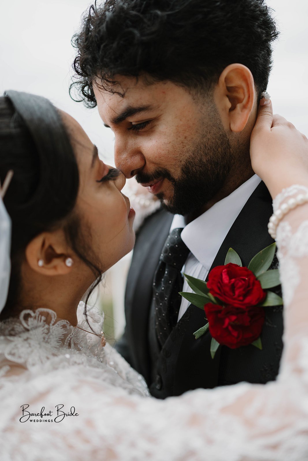 A close-up of a bride and groom sharing an intimate moment on their wedding day, with their noses touching and eyes closed. The bride wears a lace dress and pearl earrings, while the groom is dressed in a black suit with a white shirt, black tie, and