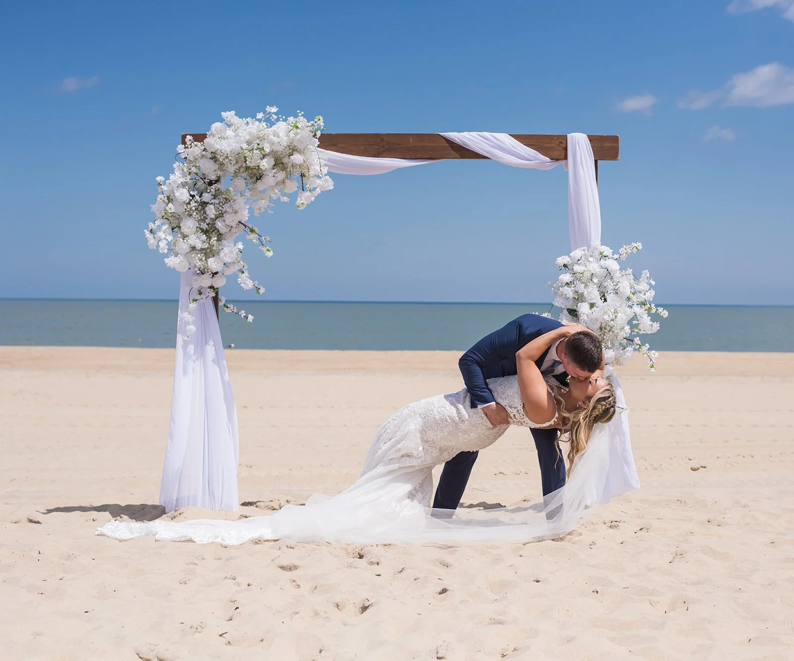 A bride and groom sharing a kiss during their beach wedding ceremony under a floral arch with the ocean in the background.