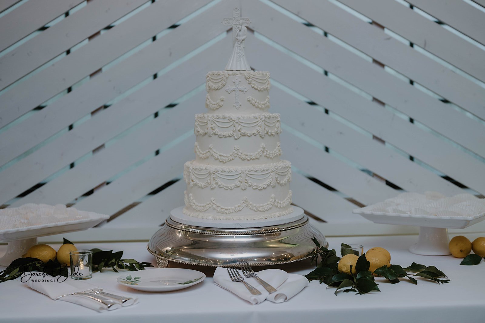 A white wedding cake with four tiers decorated with piped icing designs and a cross on the middle tier, topped with a religious figure. The cake sits on a silver cake stand on a decorated table with lemons, foliage, and plates, against a white wooden
