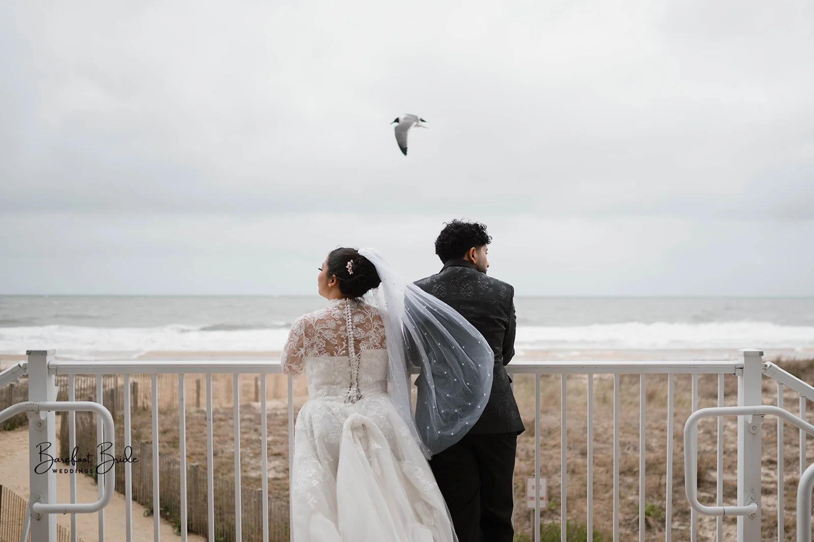 A bride and groom stand together on a balcony overlooking the ocean on a cloudy day, with a seagull flying in the sky above them.
