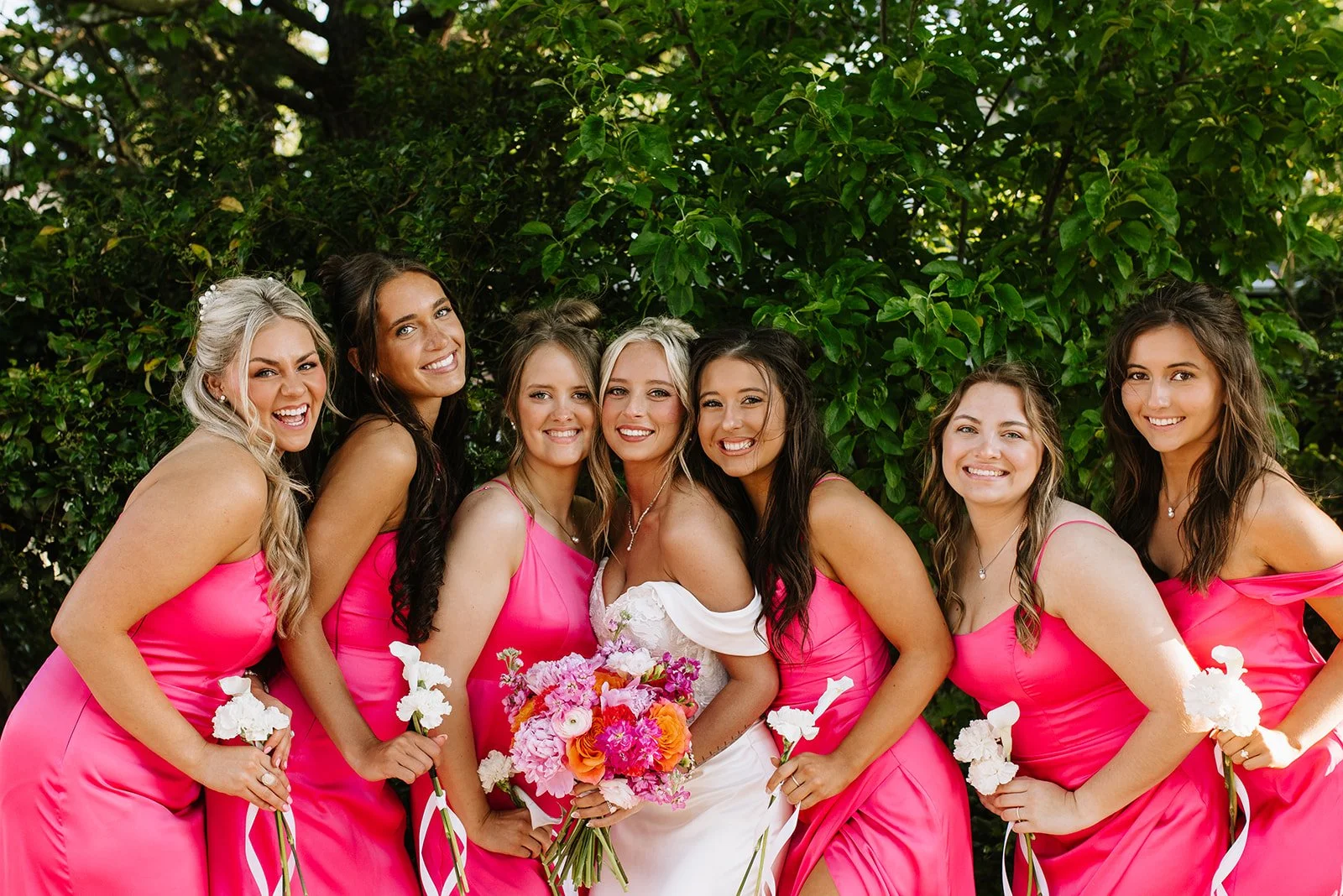 Group of six women in pink dresses and one woman in white dress holding a bouquet of flowers, posing outdoors in front of greenery.