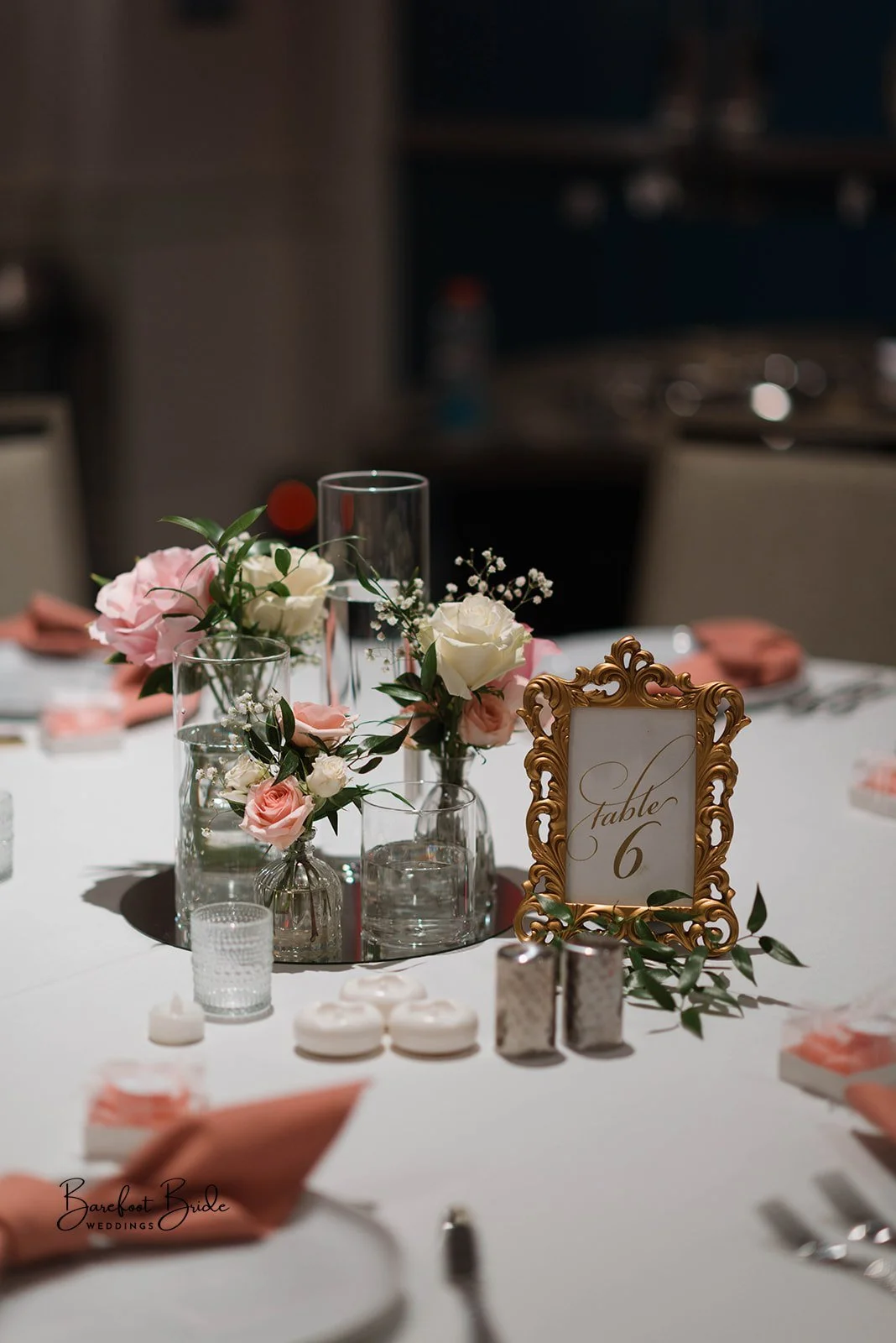Elegant wedding table centerpiece with pink and white roses, greenery, glass vases, a gold ornate table number frame displaying "table 6," and pink napkins on a white tablecloth.