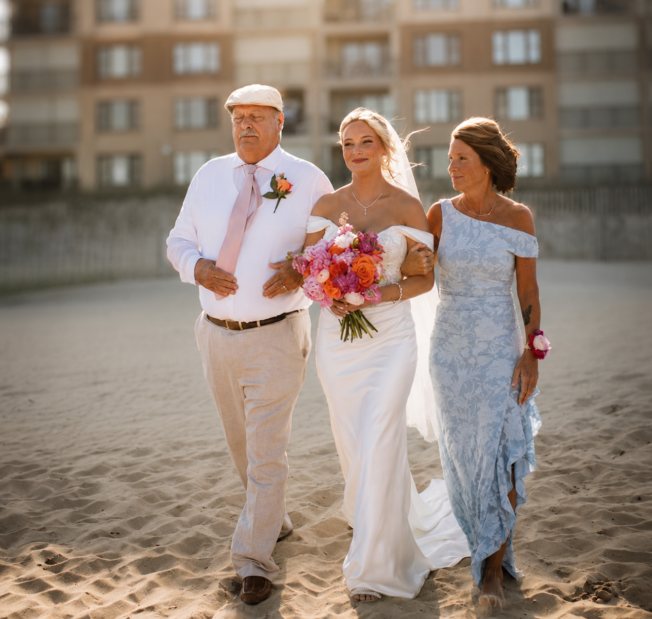 A bride in a white wedding gown being escorted on the beach by an older man and a woman, both dressed in formal attire, with a high-rise building in the background.