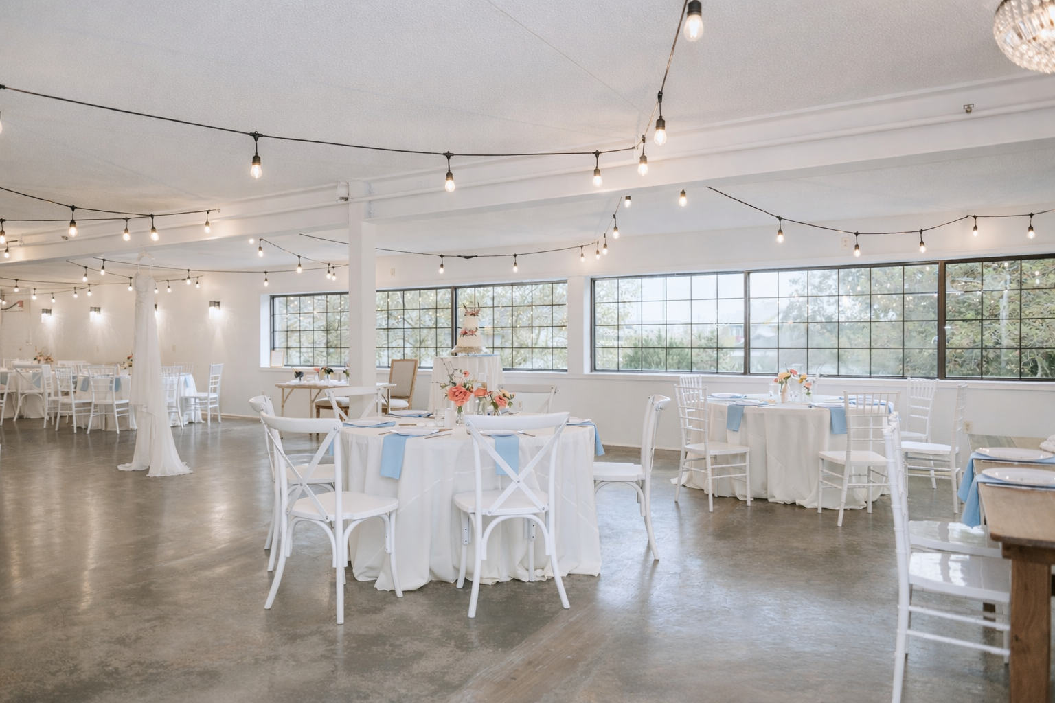 Decorated banquet hall with white tables, chairs, a wedding cake, soft lighting, and large windows overlooking trees.