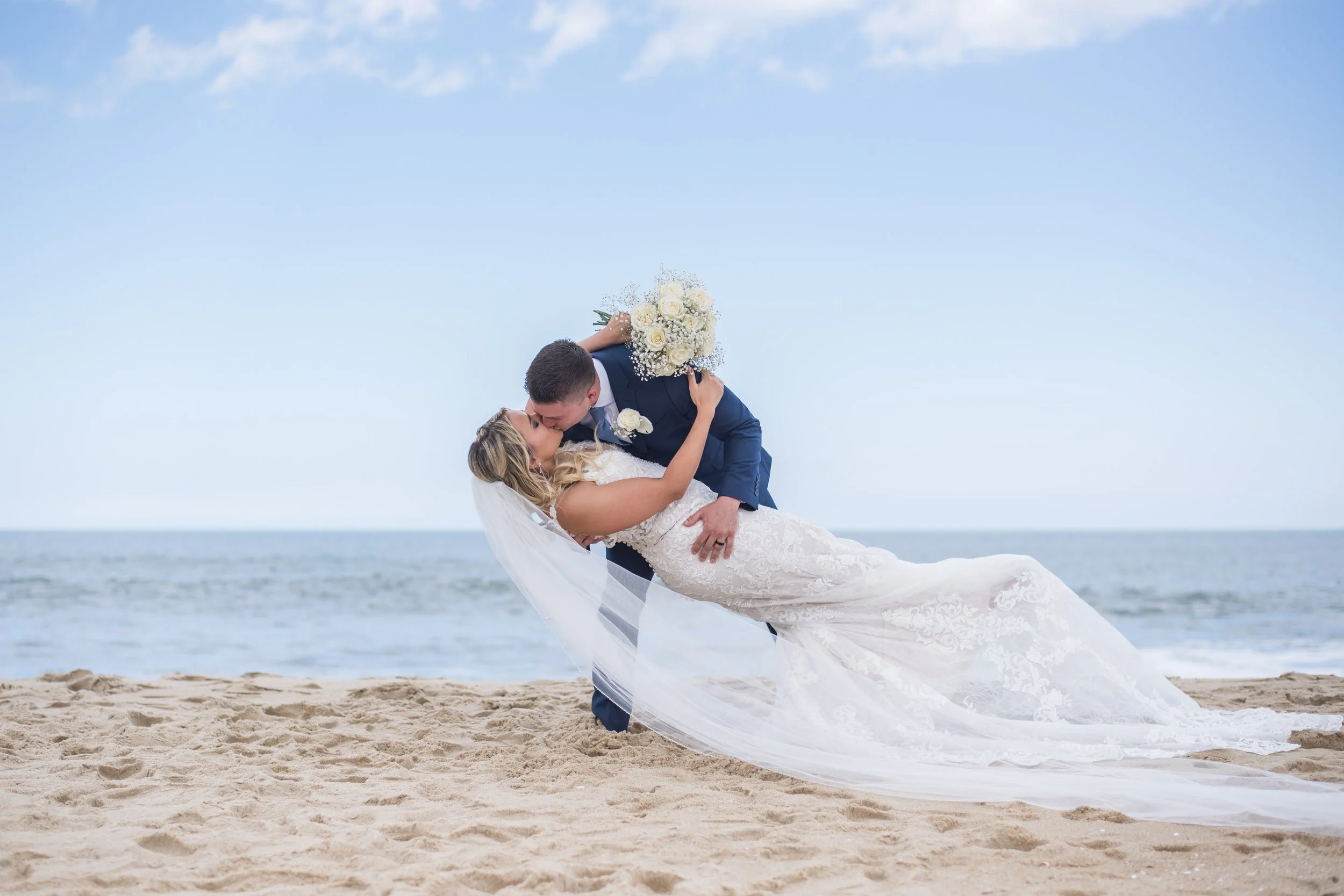 A bride and groom are on a beach, with the groom dipping the bride while they kiss. The bride holds a bouquet of white flowers, and the groom wears a navy suit. The sky is blue with some clouds, and the ocean is visible in the background.