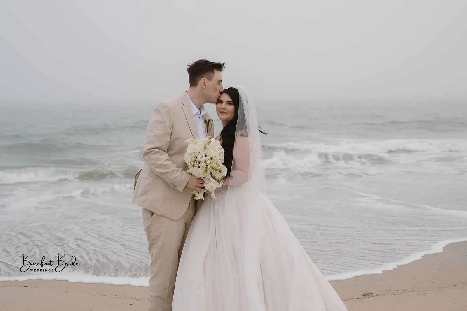 A newlywed couple standing on a beach, with waves in the background. The groom is kissing the bride on the forehead, and she is holding a bouquet of white flowers. The bride is wearing a white gown with a veil, and the groom is dressed in a tan suit.