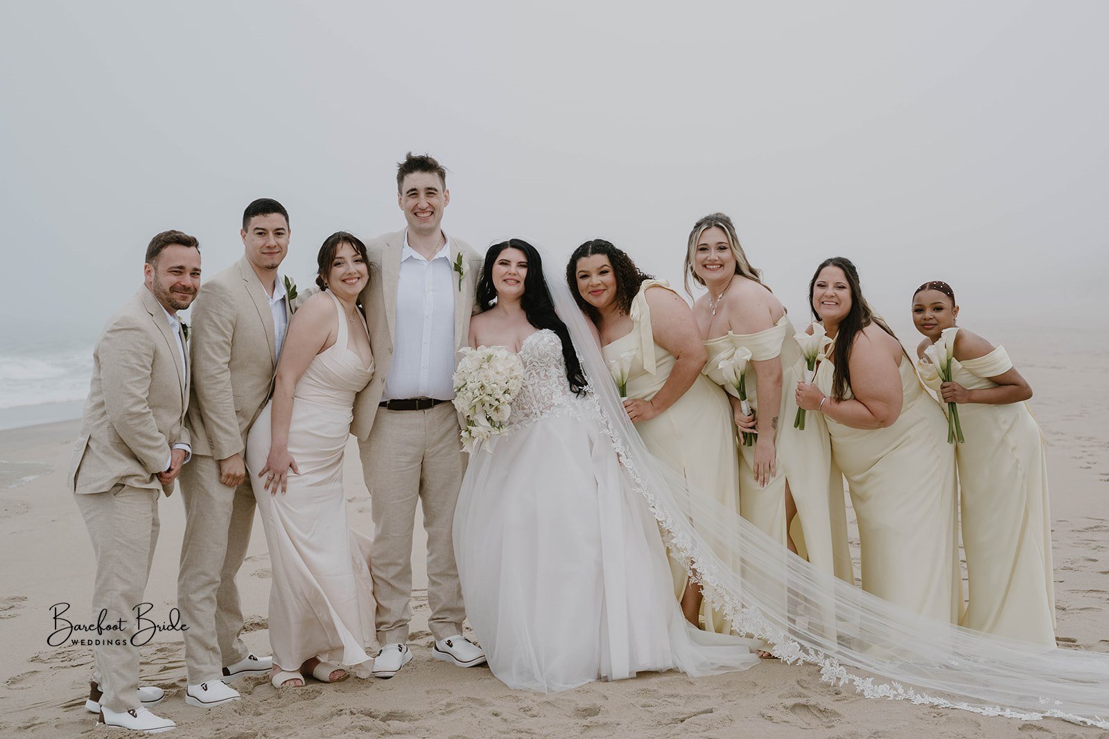 A group of people dressed in wedding attire standing on a beach with sand and ocean in the background. The bride is in a white wedding gown holding a bouquet of flowers. Bridesmaids are in matching light yellow dresses holding calla lilies, and groom