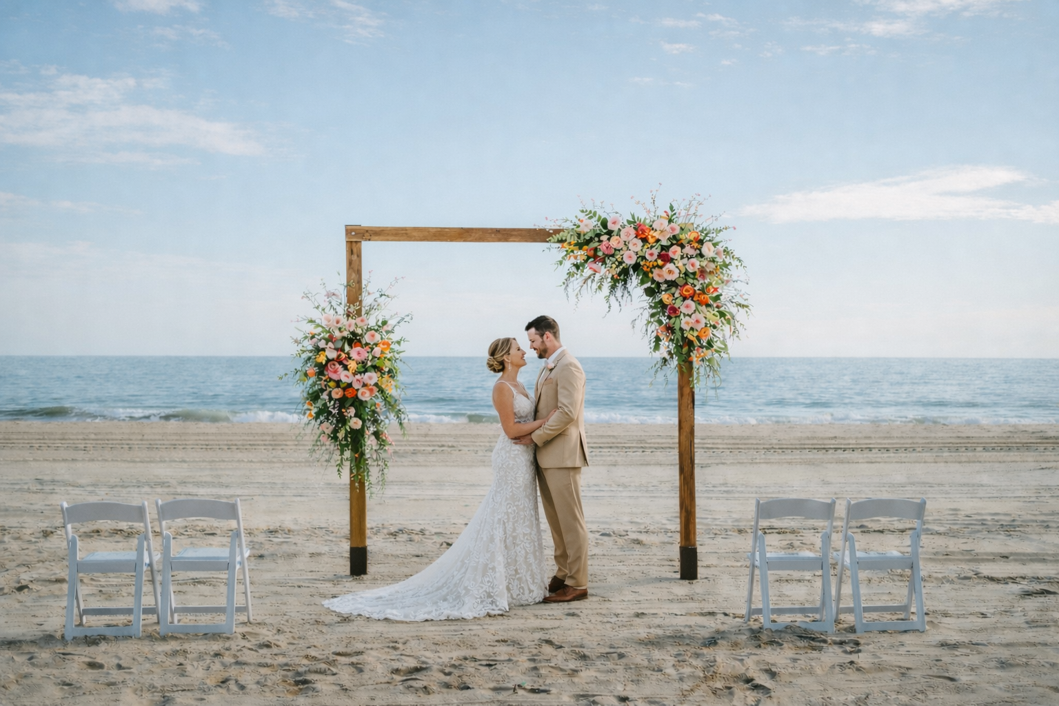 A bride and groom standing under a wooden wedding arch decorated with colorful flowers on a beach, with four empty white chairs nearby, ocean in the background.