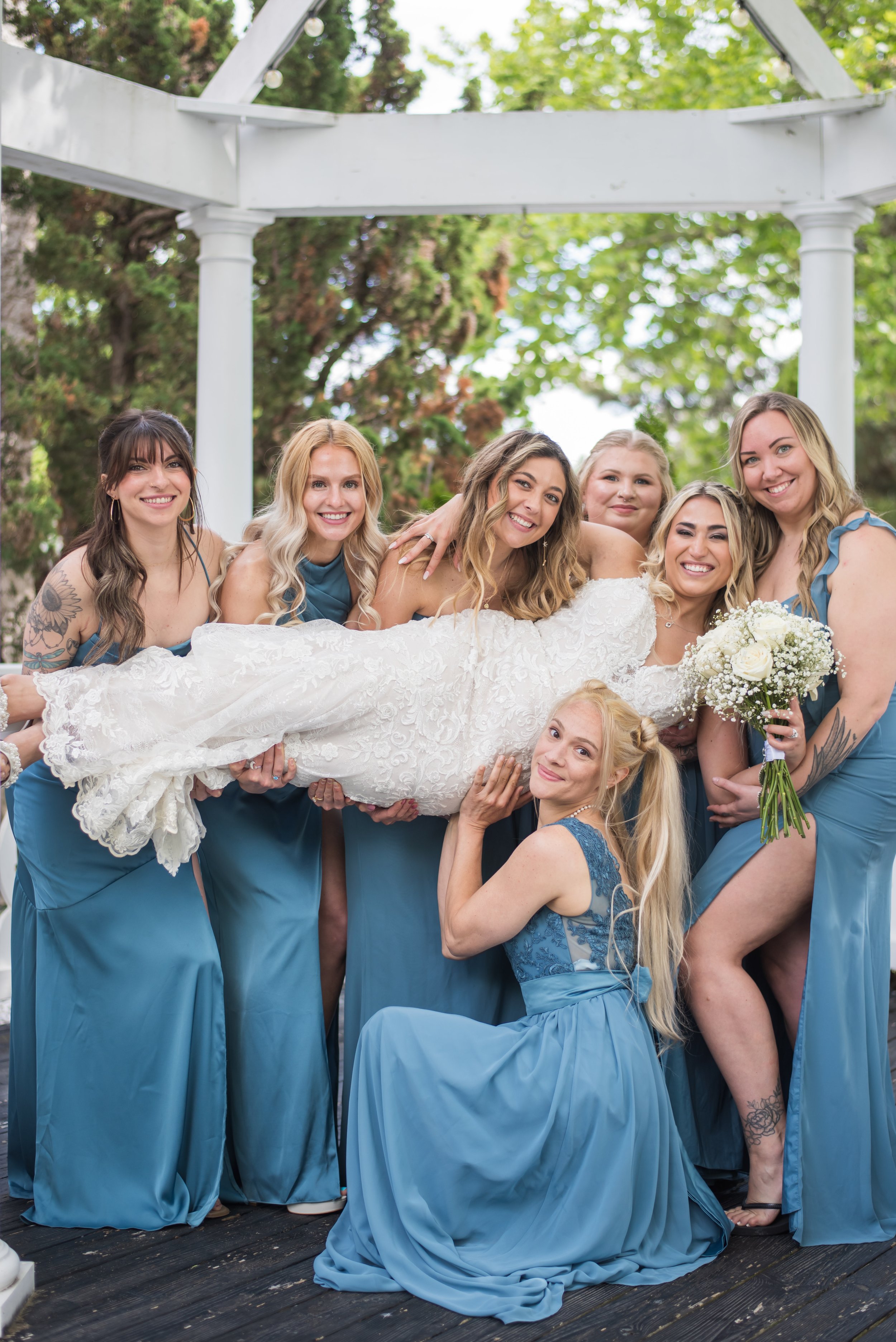 A group of women in blue dresses holding a bride in a white lace wedding dress outdoors during a wedding celebration, surrounded by trees.