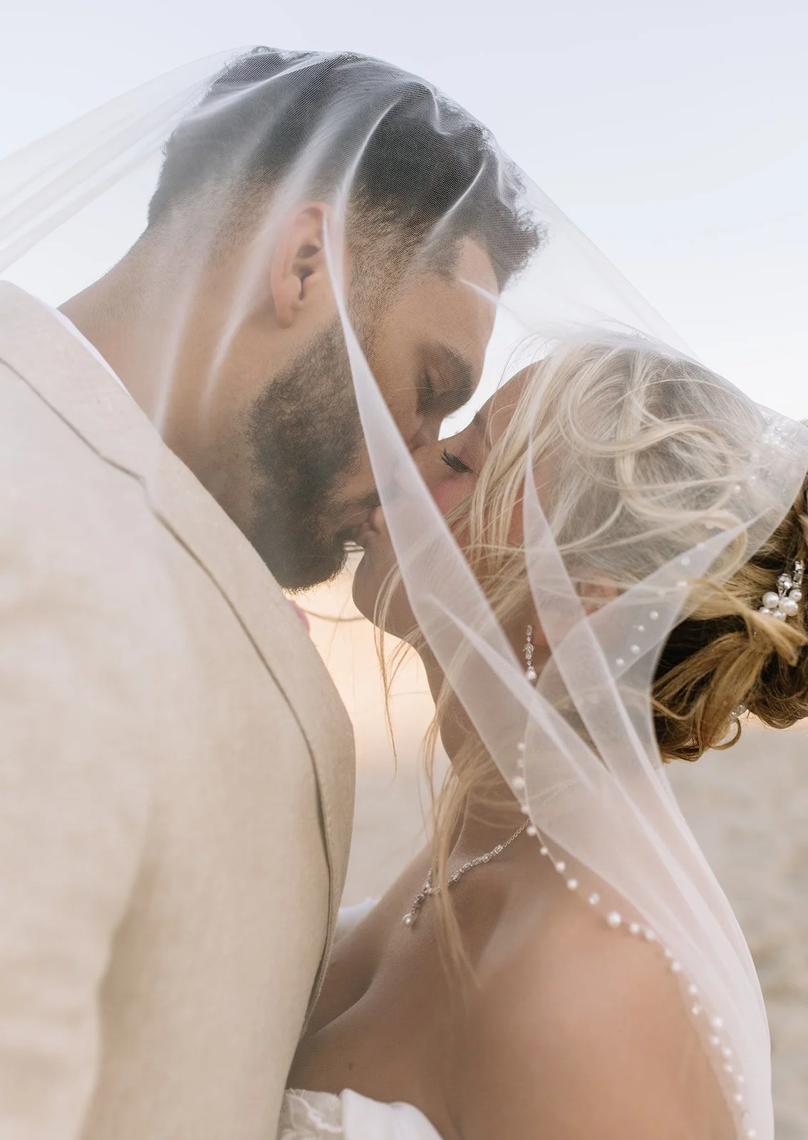 A close-up of a bride and groom sharing a kiss under a veil, with soft natural lighting, during their wedding.