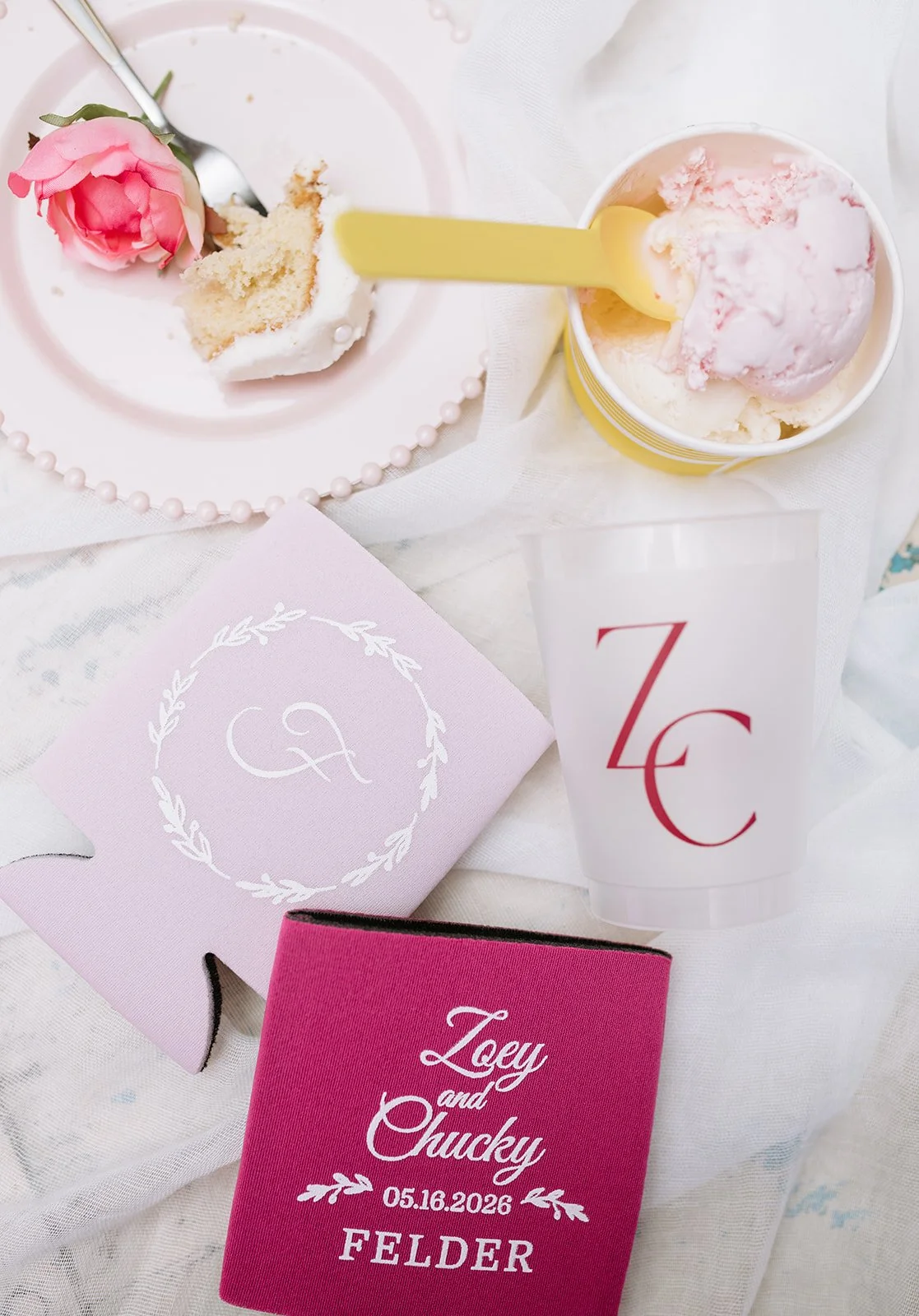 A pink rose and a slice of cake with white frosting on a white plate, a cup of pink and white ice cream with a yellow spoon, a pink invitation card with initials, a white and pink glass with red initials, and a pink folder with wedding details on a w