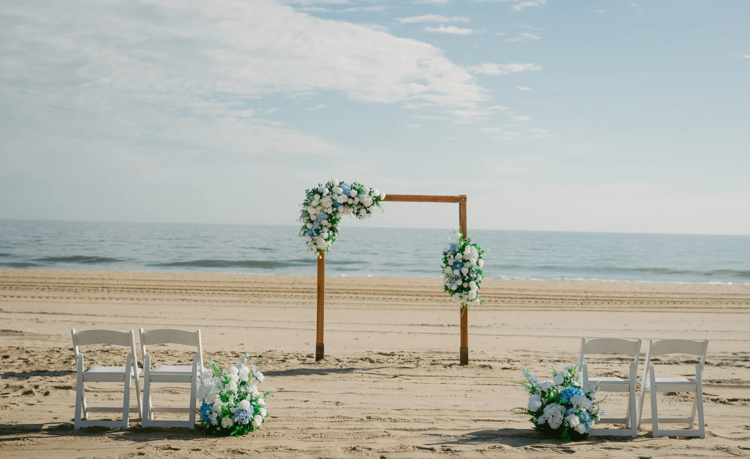 Beach wedding altar with floral decorations, white chairs, and ocean backdrop.