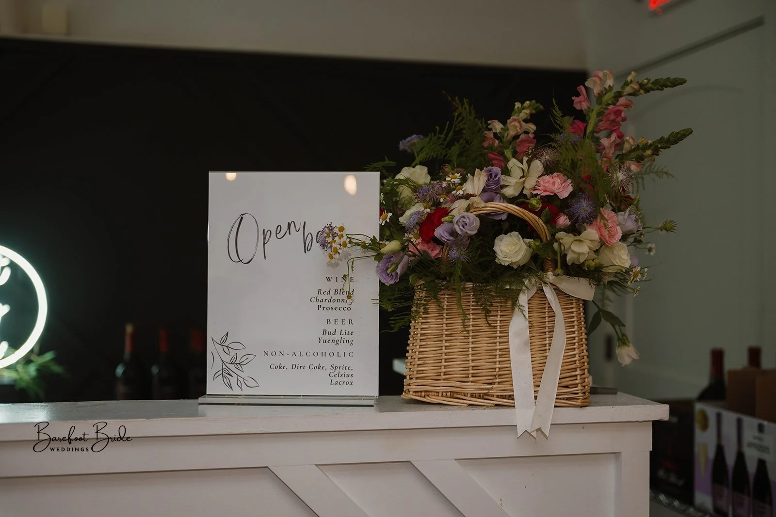 A floral arrangement in a wicker basket with pink, purple, and white flowers, next to a white sign with handwritten style text that says "Open" and a menu listing wine, beer, and non-alcoholic drinks, on a white counter with the logo "Barefoot Bride 