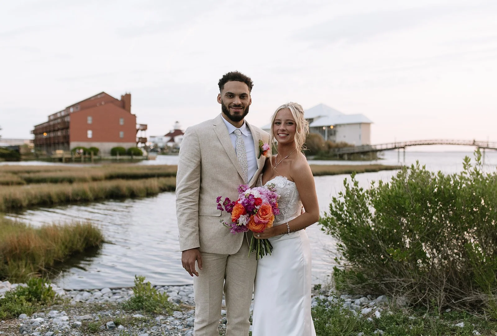 A newlywed couple standing by a river, with a man in a beige suit and a woman in a white wedding dress holding a bouquet of flowers, during sunset with buildings and a bridge in the background.